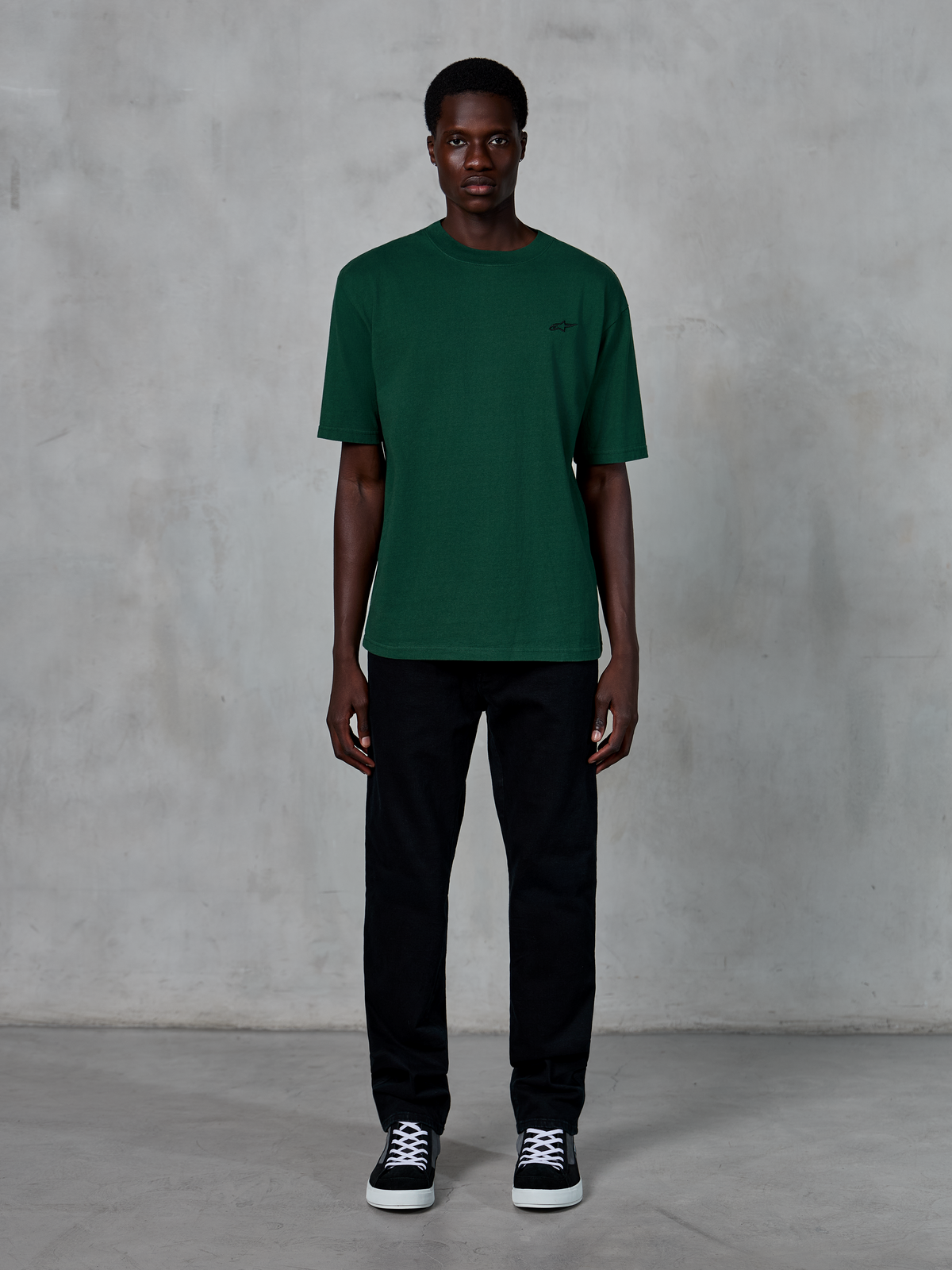 Alpinestars Essentials Ageless SS Tee, koszulka z krótkim rękawem, kolor Pine Green, z subtelnym czarnym logo na piersi, noszona przez modela w czarnych spodniach i sneakersach, casualowy styl streetwear o luźnym kroju