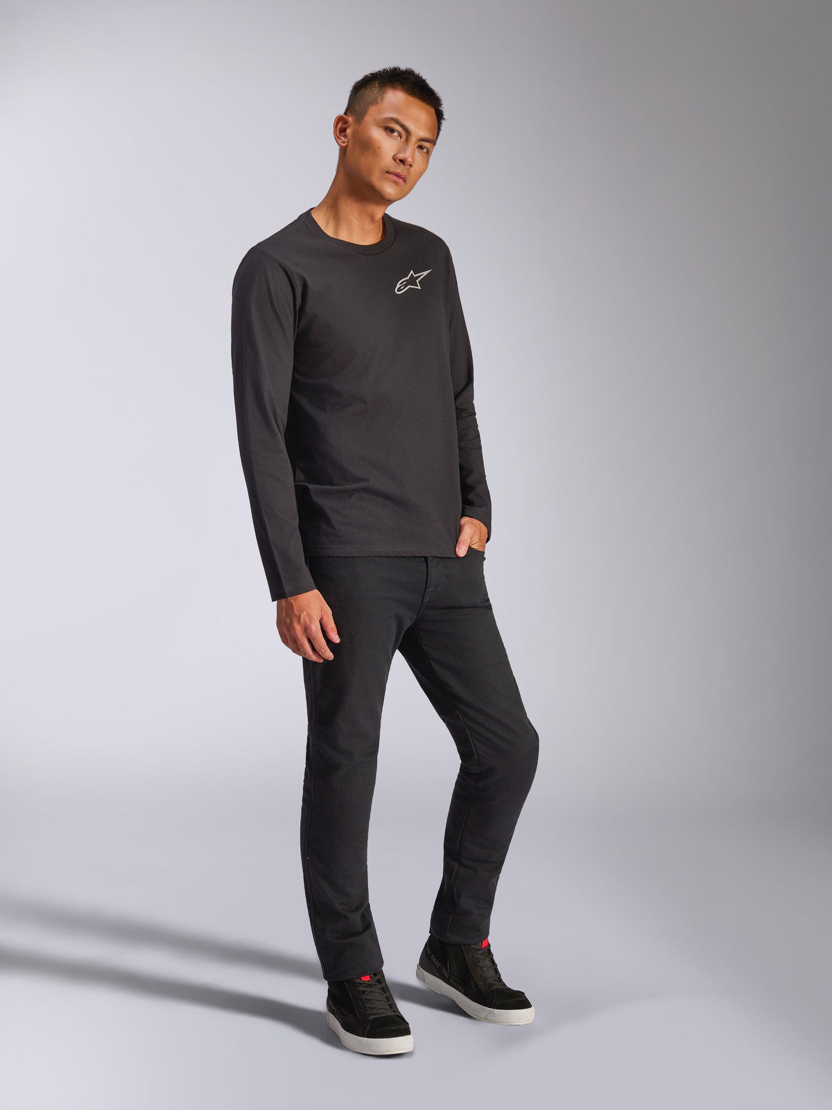 Alpinestars Trackside CSF Tee, koszulka z długim rękawem, czarna, białe logo Astars na piersi, noszona przez modela z czarnymi spodniami i sneakersami, casualowy streetwear inspirowany motorsportem