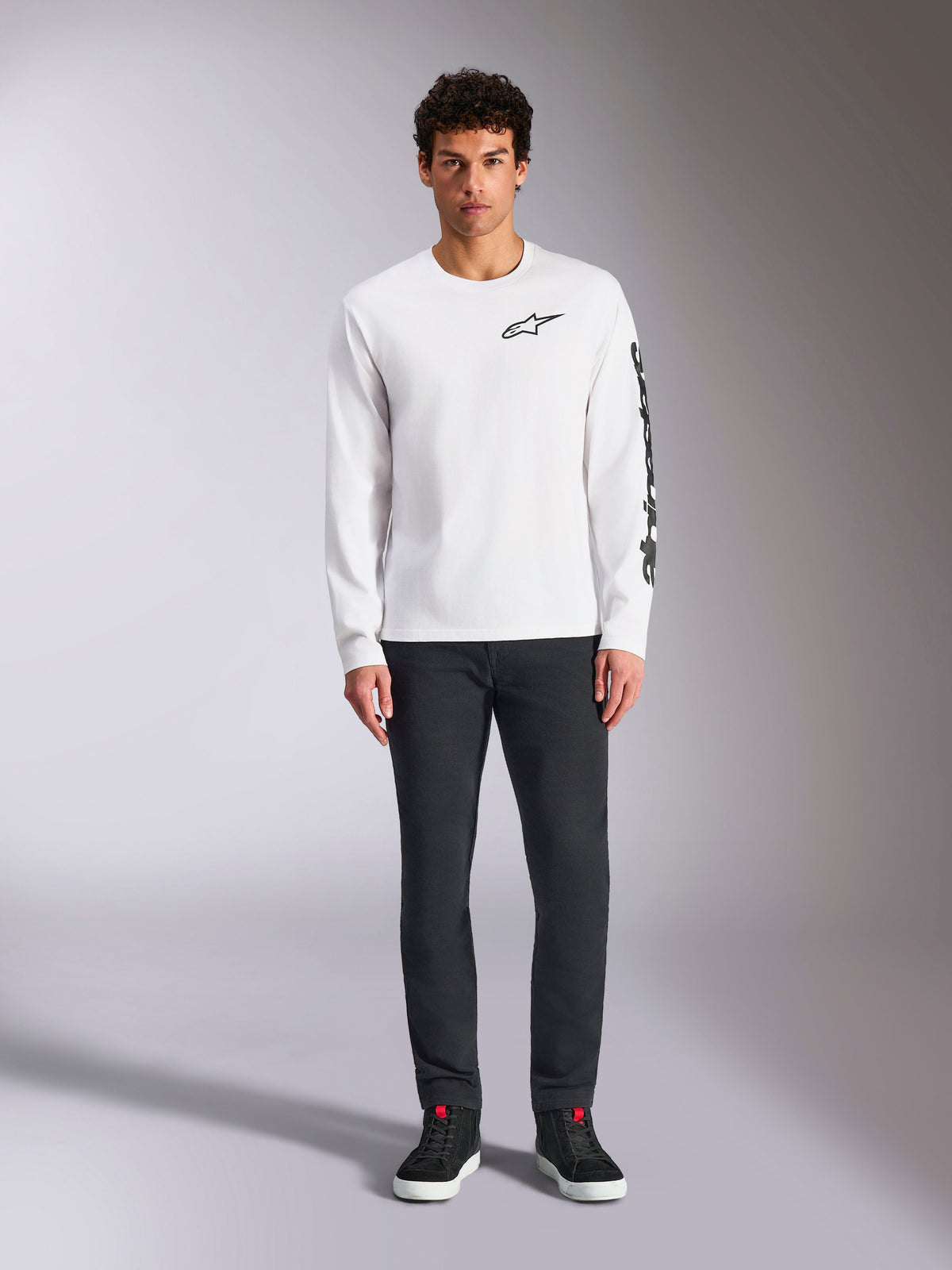 Alpinestars Trackside CSF Tee - Long Sleeve, biała z czarnym brandingiem, małe logo na piersi i pionowy napis na rękawie, prezentowana przez modela w ciemnych spodniach i czarnych sneakersach, streetwear inspirowany sportami motorowymi