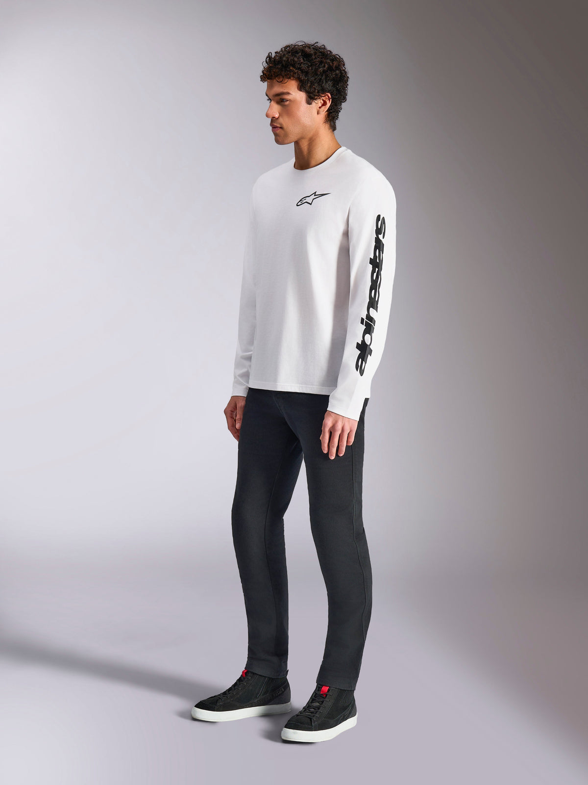 Alpinestars Trackside CSF Tee, biała koszulka z długim rękawem, czarne logo z gwiazdą na piersi i napis na lewym rękawie, model w ciemnych spodniach i sneakersach, sportowa odzież lifestyle