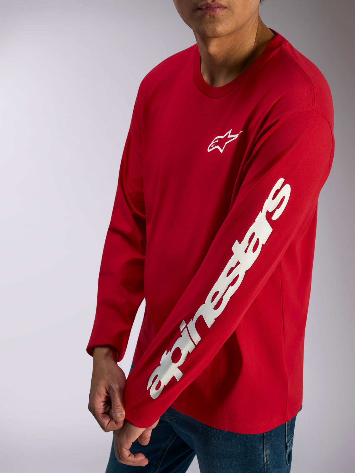 Alpinestars Trackside CSF Tee, koszulka z długim rękawem, czerwona, białe logo Alpinestars na piersi i wyraźny napis na rękawie, codzienna odzież streetwearowa w stylu motorsportowym noszona z niebieskimi jeansami