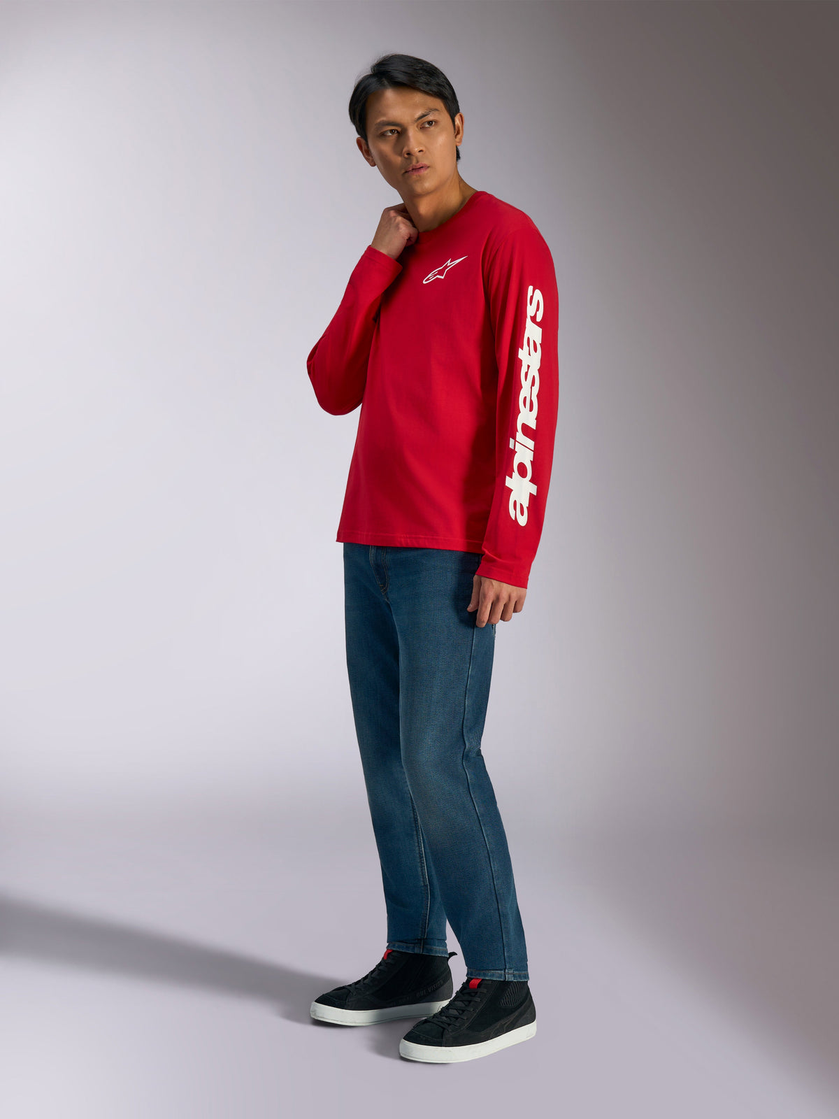 Alpinestars Trackside CSF Long Sleeve Tee, czerwona koszulka z długim rękawem z białym logowaniem Alpinestars na klatce piersiowej i rękawie, noszona przez modela z niebieskimi dżinsami i czarnymi sneakersami, codzienna odzież uliczna inspirowana motorsportem