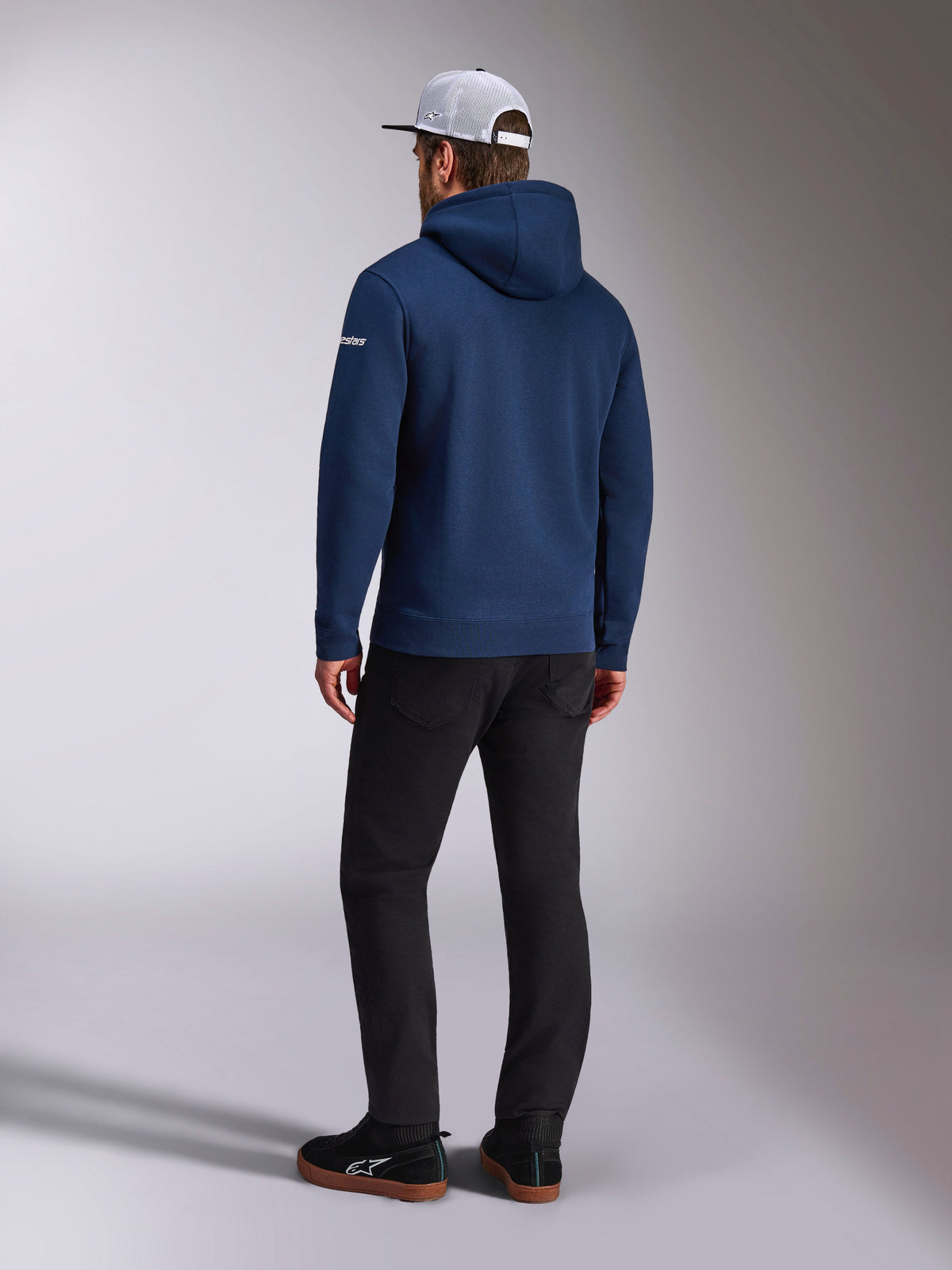 Alpinestars Frontal Hoodie, Rear view of a navy blue pullover fleece hoodie, white word logo on the sleeve, paired with black pants and a white mesh trucker hat, casual motorsport-inspired streetwear