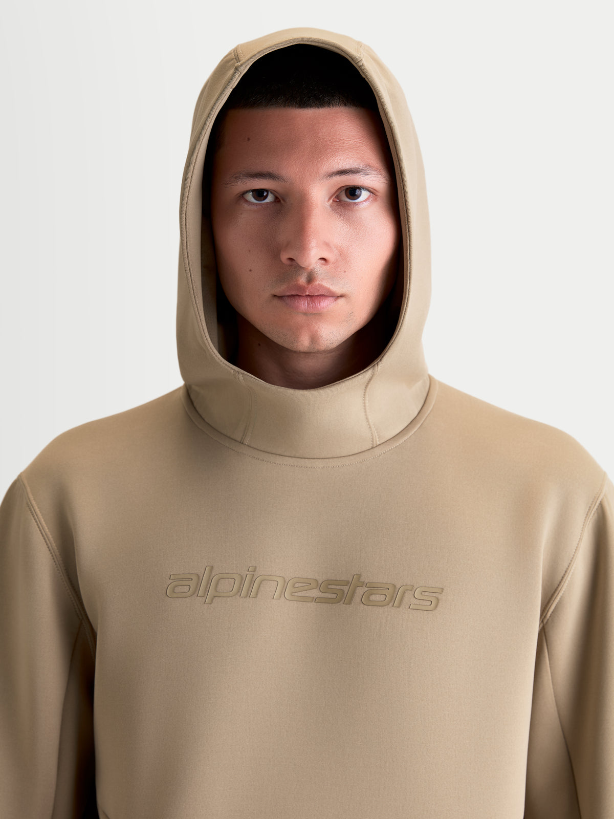 Alpinestars Datrix Hoodie, brązowa bluza z kapturem typu pullover, odzież sportowa unisex, zbliżenie na modela z założonym kapturem, tonalne logo Alpinestars na piersi, materiał polarowy premium z mieszanki trzech włókien, styl casualowy.