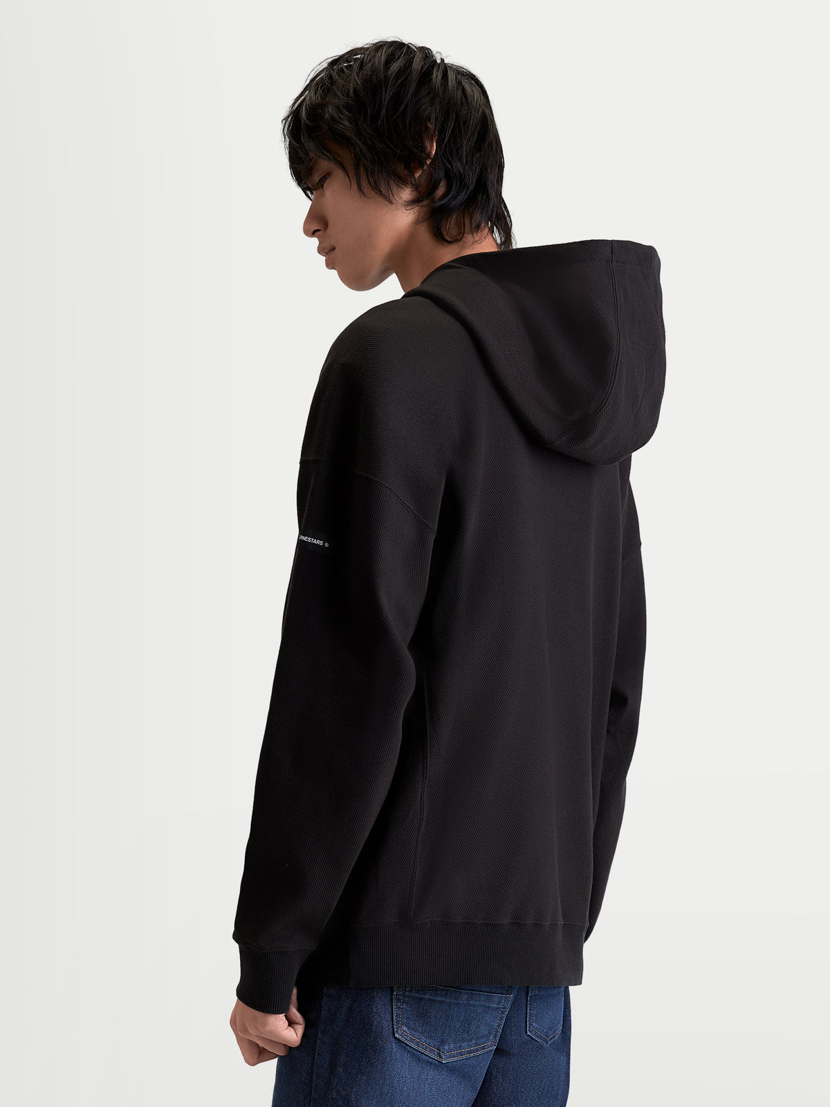 Alpinestars bluza z kapturem Camino Textured Oversized, męska bluza typu pullover, czarna, teksturowany polar o splocie twill, wysoki kołnierz, widok z tyłu ukazujący luźną sylwetkę i tkaną metkę na rękawie.