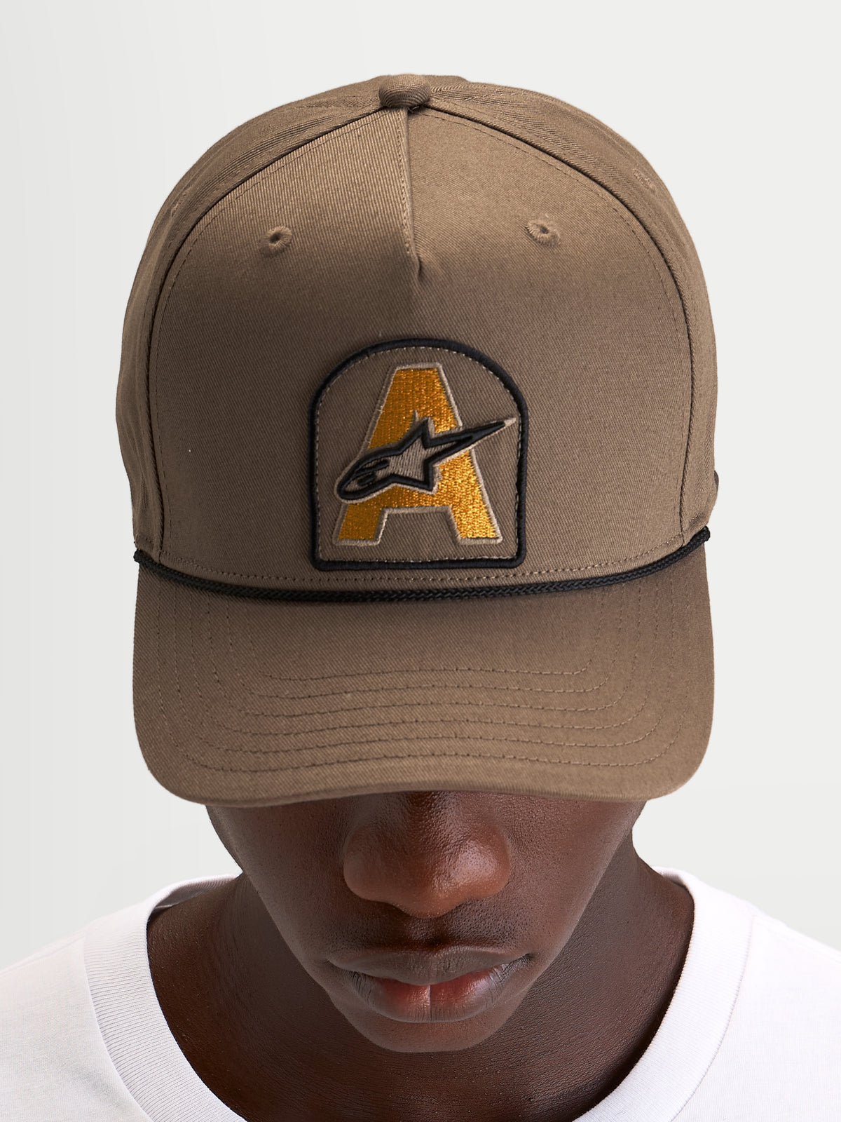 Alpinestars Czapka Acme Snapback, brązowa czapka z daszkiem typu sportswear z naszywką z haftowanym logo Astars i czarnym sznurkiem, prezentowana na modelu