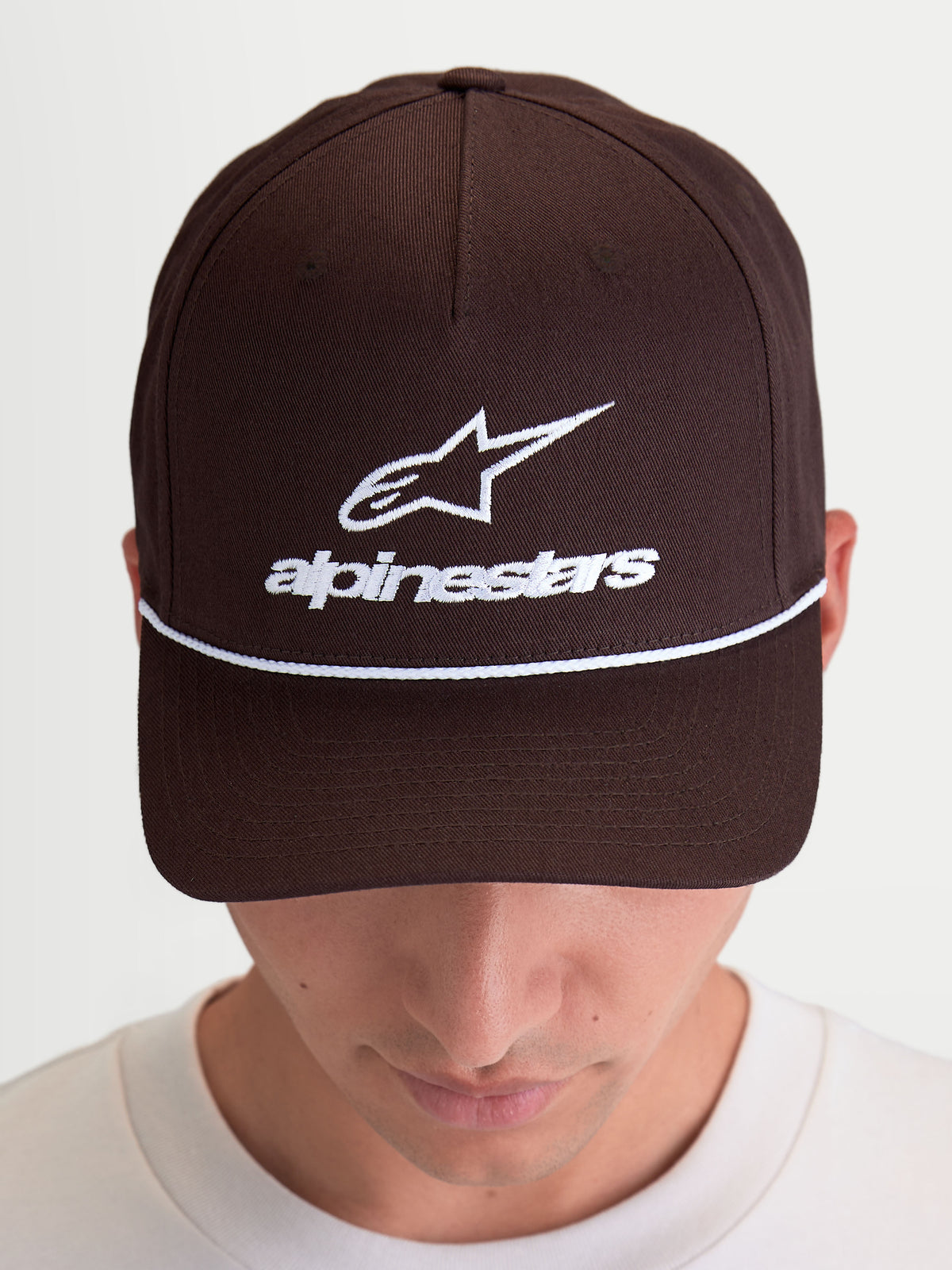 Alpinestars Czapka Combo Logo Snapback, brązowa czapka z daszkiem typu sportswear, biały haft logo Astars i logotyp z przodu, zakrzywiony daszek, usztywniona korona, pokazana na modelu.
