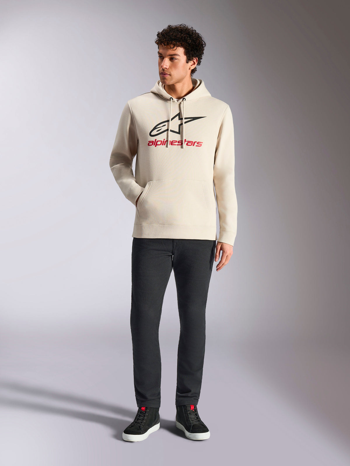 Alpinestars Always V3 Hoodie, casualowa odzież streetwear, kolor złamanej bieli z czarnym logo gwiazdy i czerwonym napisem, kaptur ze sznurkiem, długie rękawy, noszona przez modela z czarnymi spodniami i czarnymi sneakersami
