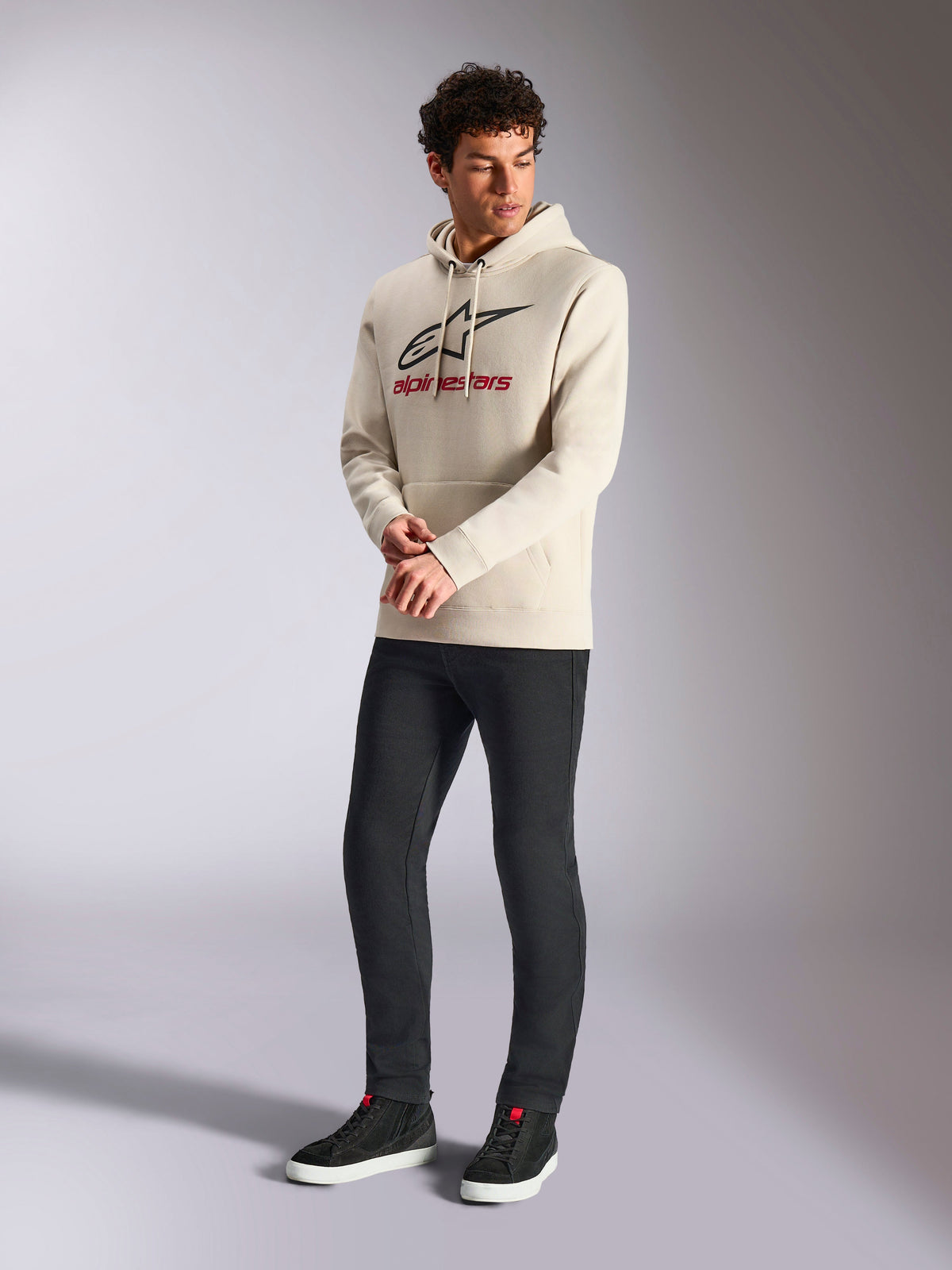 Alpinestars Always V3 Hoodie, polarowa bluza z kapturem typu pullover w kolorze złamanej bieli, z przodu linearne logo z czarną gwiazdą i czerwonym brandingiem, noszona przez modela z czarnymi spodniami i czarnymi sneakersami, casualowy styl motorsportowy
