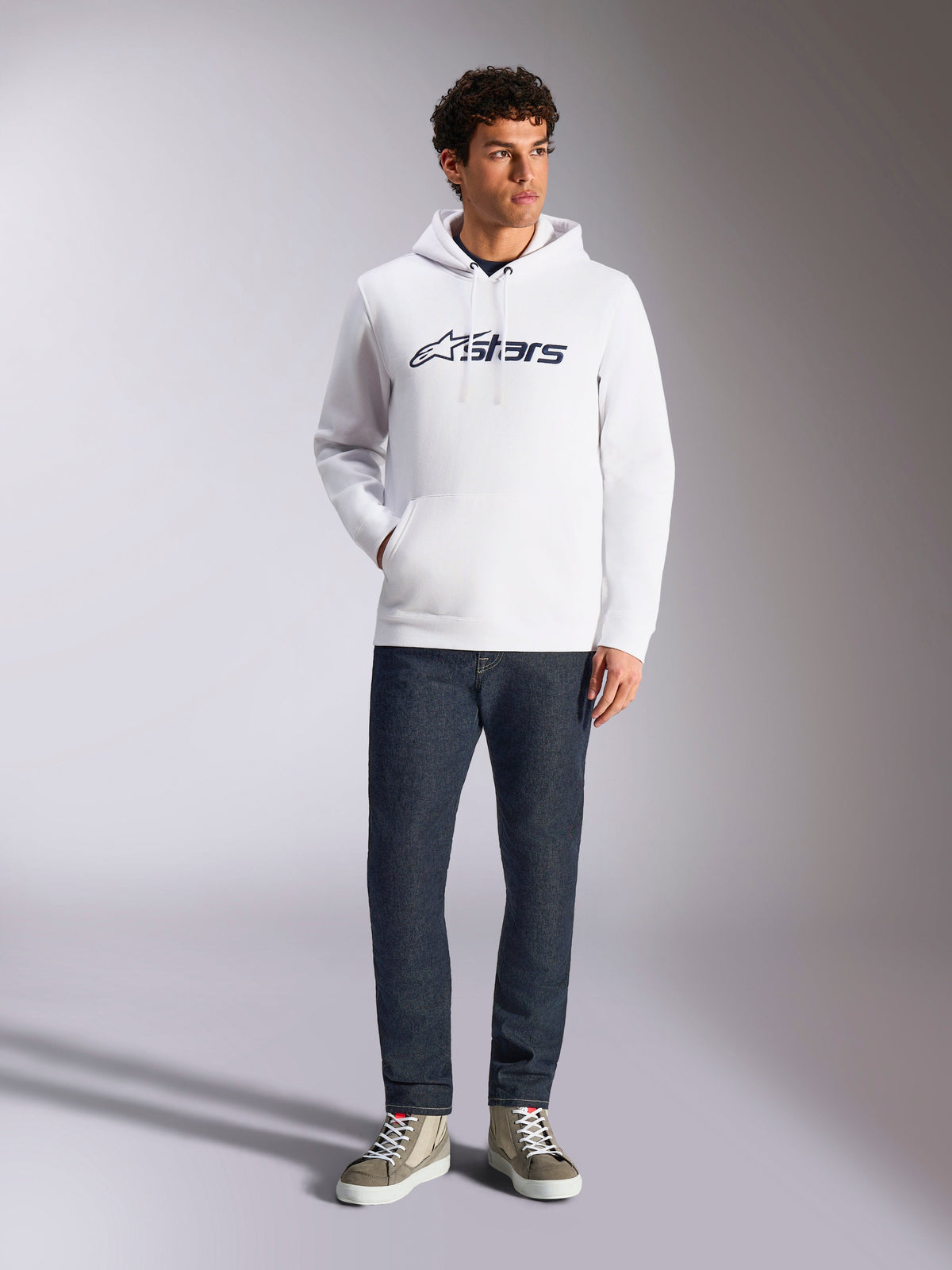 Alpinestars Blaze V3 Hoodie, white casual streetwear hoodie with navy Alpinestars logo on the chest, worn by a model with dark blue jeans and tan sneakers, featuring a drawstring hood and front pouch pocket
