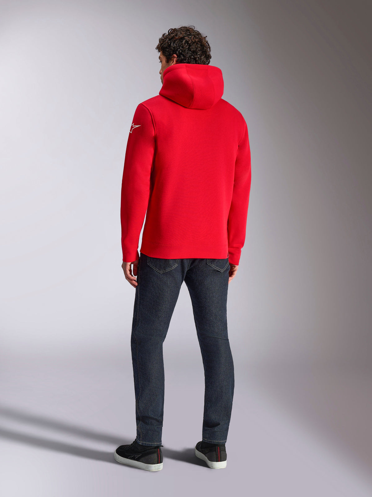 Alpinestars Linear Wordmark V3 Hoodie, casual red pullover hoodie, back view featuring the hood and white Ageless logo on the sleeve, paired with dark blue jeans and black shoes, lifestyle motorsport streetwear