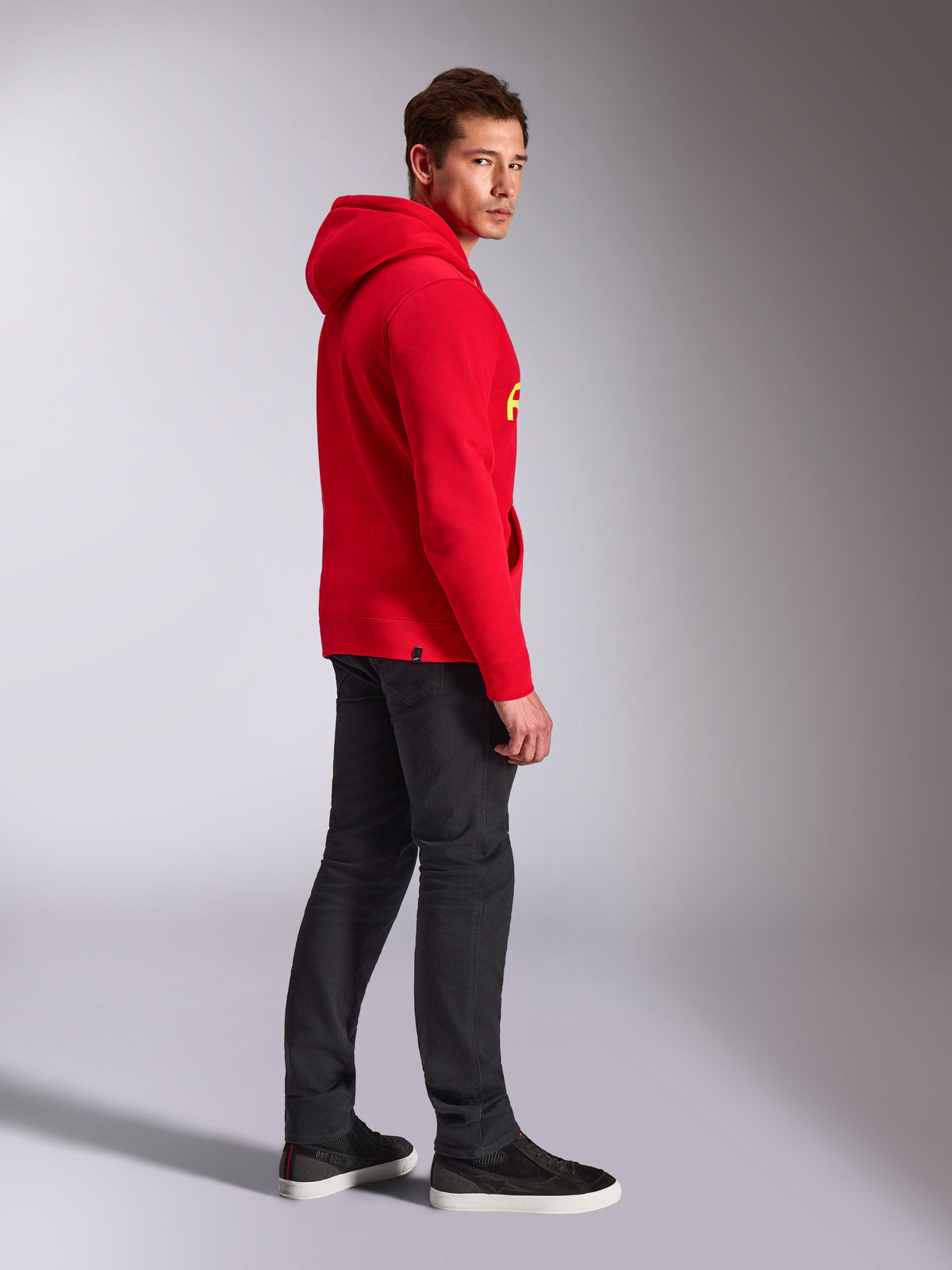 Alpinestars Linear Wordmark V3 Hoodie, Red, casual streetwear, model viewed from the side and back wearing a red pullover hoodie with yellow branding, dark grey jeans, and black sneakers, studio lifestyle shot