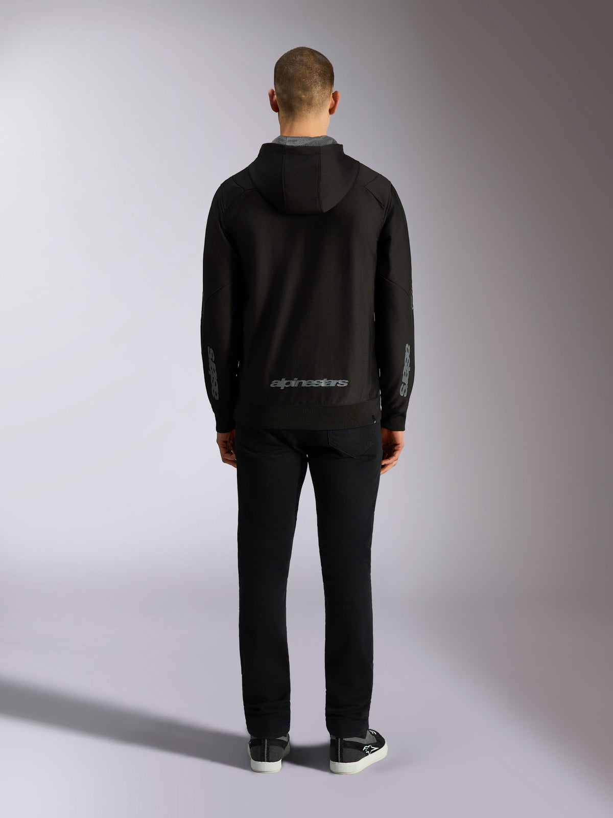 Alpinestars Sessions V3 Hoodie, czarno-szara bluza z kapturem, widok z tyłu, szary napis Alpinestars na dole pleców i logo astars na przedramionach, model w czarnych spodniach, codzienna odzież streetwearowa inspirowana sportami motorowymi