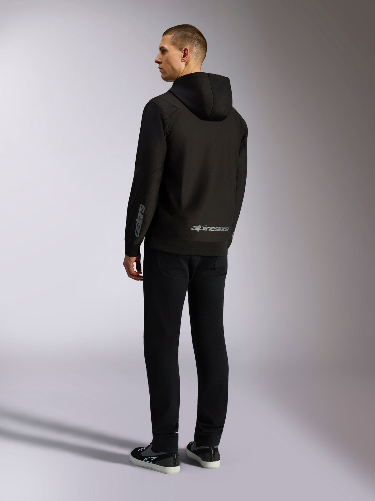 Alpinestars Sessions V3 Hoodie, czarna bluza streetwearowa, widok od tyłu na modelu, szary napis Alpinestars na dole pleców i logo Astars na rękawie, fason z kapturem zapinany na zamek, nowoczesny styl inspirowany motorsportem