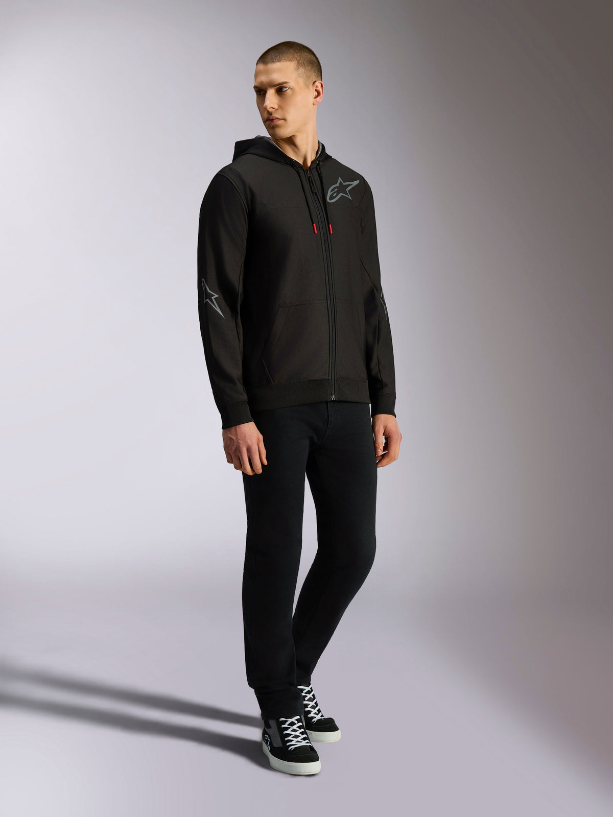 Alpinestars Sessions V3 Hoodie, czarno-szara polarowa bluza z zamkiem z przodu, szare logo na piersi i rękawach, kaptur ze sznurkami o czerwonych końcówkach, widok na całą sylwetkę modela w codziennym stroju streetwearowym inspirowanym motorsportem