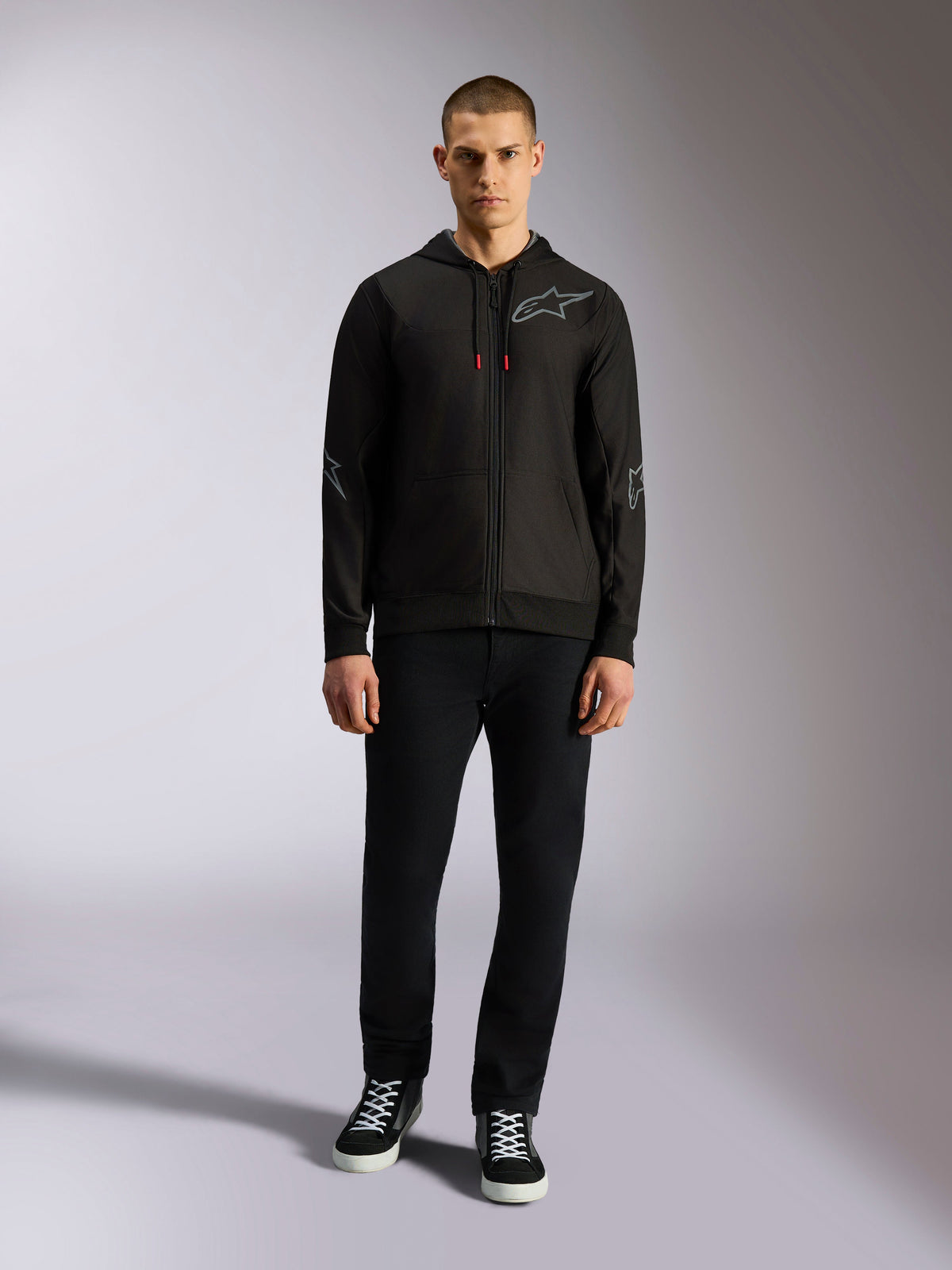 Alpinestars Sessions V3 Hoodie, czarna rozpinana bluza z kapturem z szarym logo Astars na piersi, grafikami na łokciach i przedramionach, czerwonymi akcentami na sznurkach, noszona przez modela w czarnych spodniach i sneakersach, codzienna odzież motorsportowa