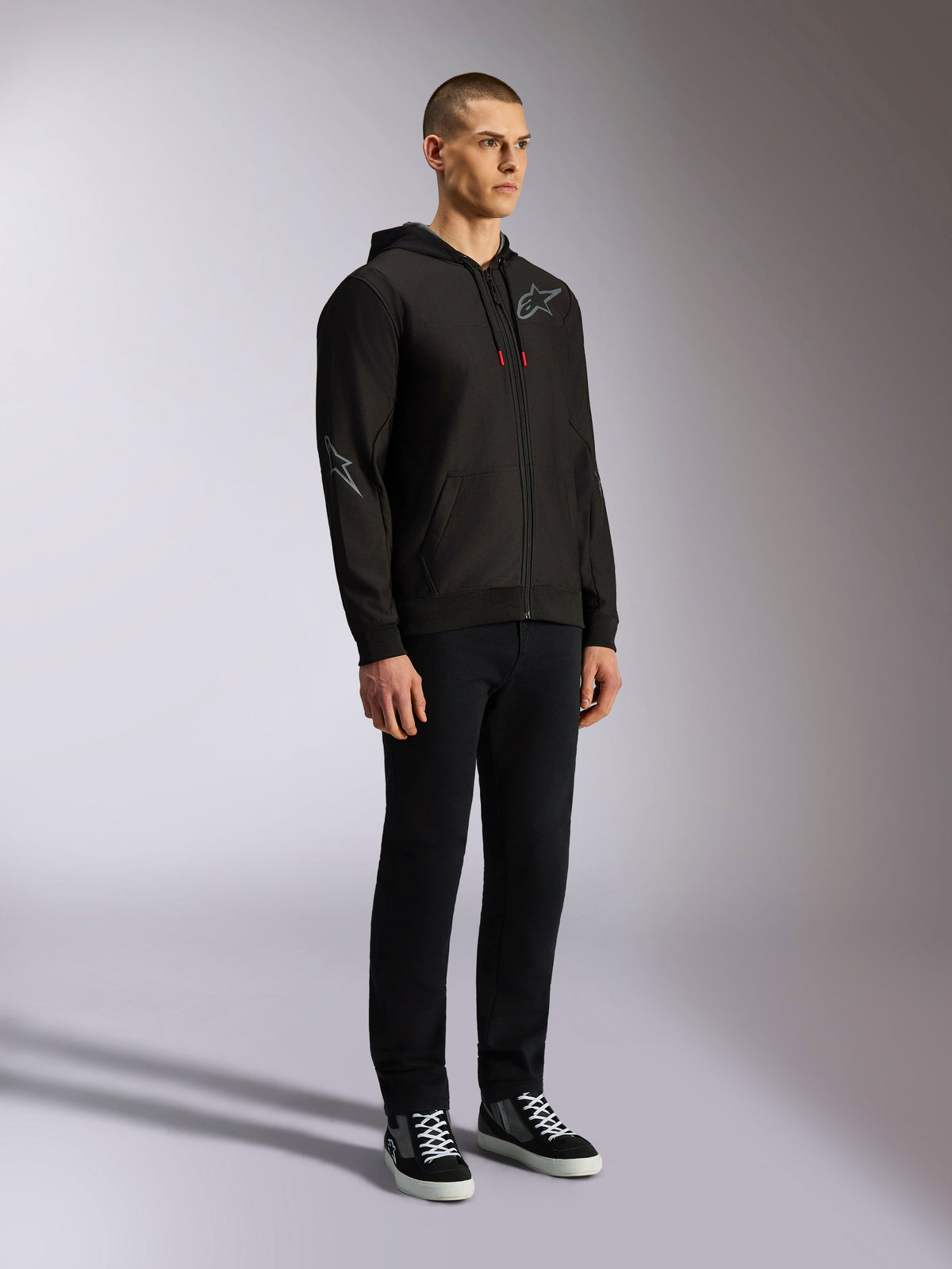 Bluza z kapturem Alpinestars Sessions V3, codzienna odzież streetwearowa w stylu motorsportowym, czarna bluza rozpinana z szarym logo gwiazdy na piersi i rękawie, sznurki z czerwonymi końcówkami, ściągacze na mankietach i dole, noszona przez modela z czarnymi spodniami i sneakersami.