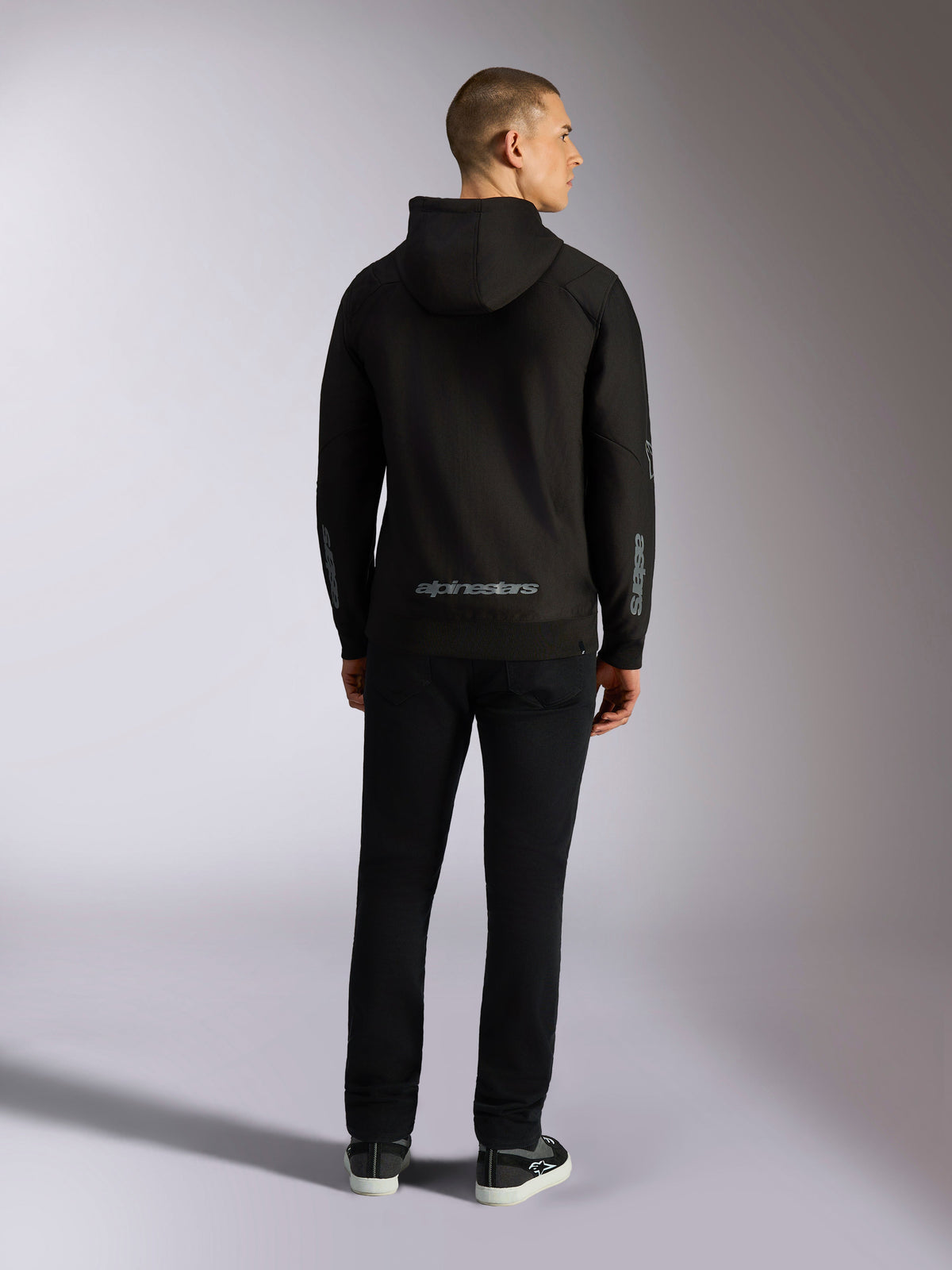 Bluza z kapturem Alpinestars Sessions V3, casualowa odzież uliczna, czarna z szarym napisem Alpinestars na dole pleców i brandingiem „astars” na rękawach, widok z tyłu na męskiego modelu w bluzie i czarnych spodniach, nowoczesny design inspirowany sportami motorowymi