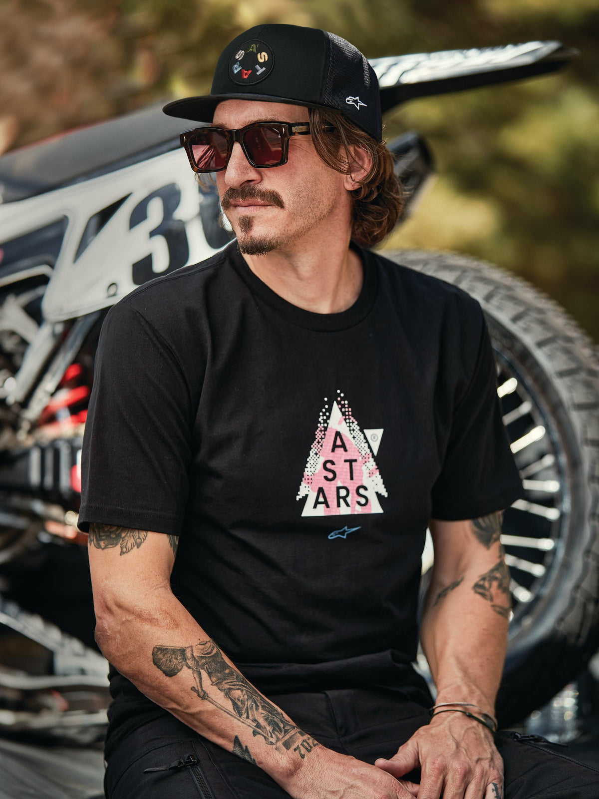 Alpinestars Gambit Trucker Hat in black with circular logo patch, worn with Alpinestars Soloist SS Tee in black, lifestyle shot featuring a man with sunglasses and tattoos sitting in front of a motocross bike.