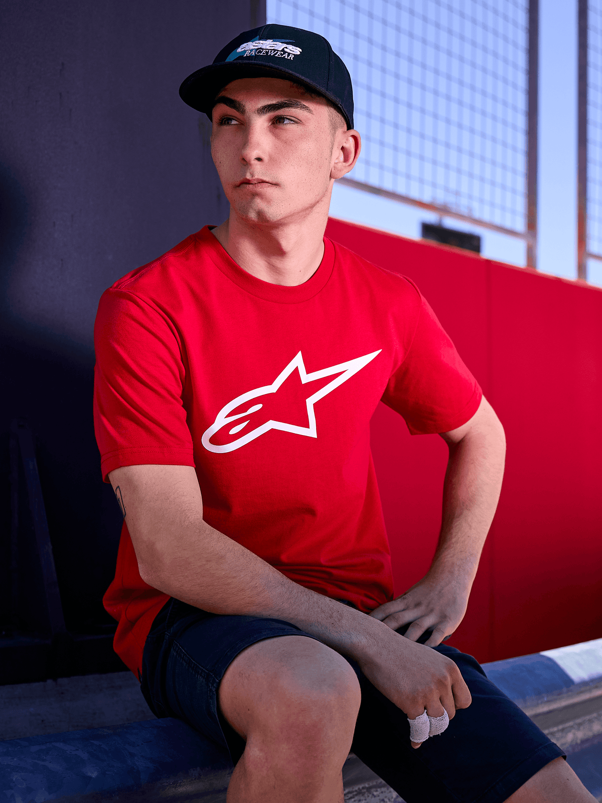 Czapka Alpinestars Profile, czarna, zdjęcie typu lifestyle przedstawiające mężczyznę w czapce z zakrzywionym daszkiem i czerwonym t-shircie z białym logo Alpinestars, codzienna odzież uliczna inspirowana sportami motorowymi