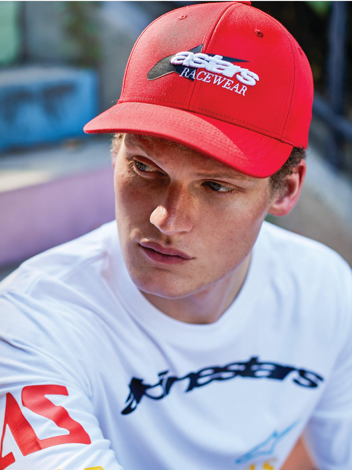 Mężczyzna w czapce Alpinestars Profile Hat, czerwona czapka z zakrzywionym daszkiem typu snapback, biało-czarny haft 'astars RACEWEAR' z przodu, biała koszulka Alpinestars z długim rękawem, motorsportowy styl lifestyle