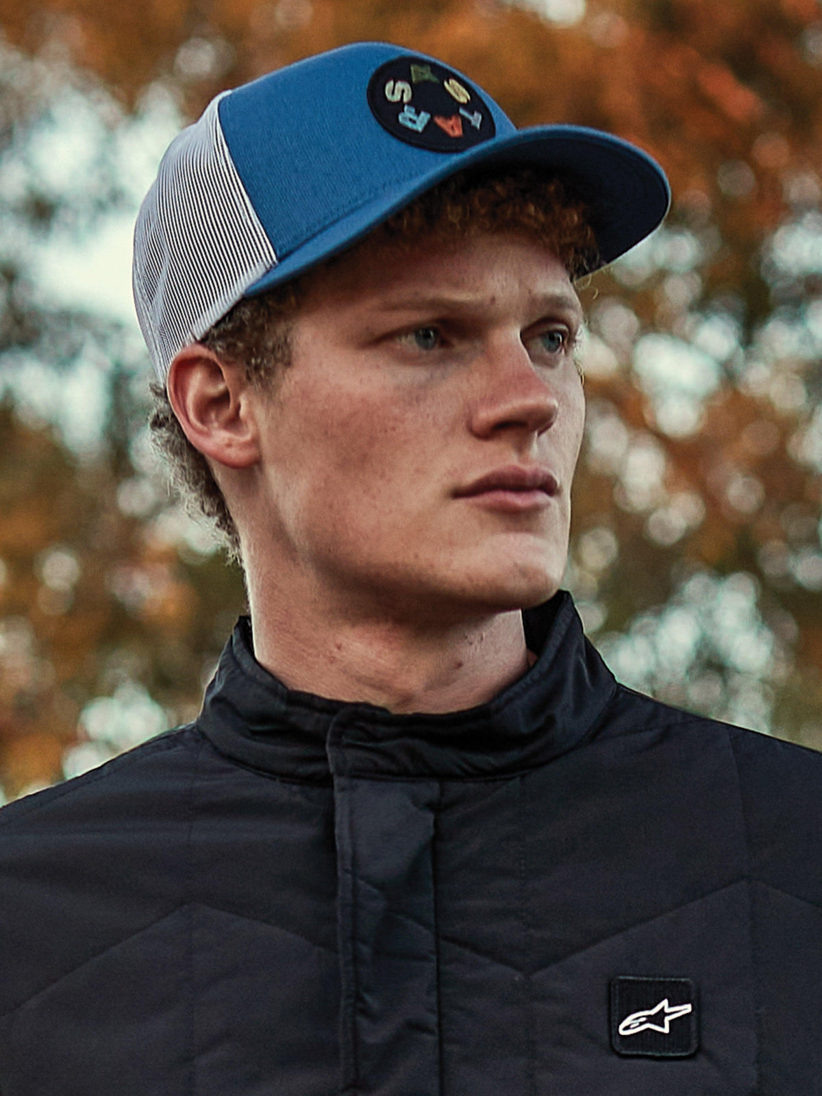 Mężczyzna w czapce Alpinestars Gambit Trucker, niebiesko-biała czapka z daszkiem i siatką z tyłu z okrągłą naszywką z logo Astars, zestawiona z czarną pikowaną kurtką Alpinestars, lifestylowy portret w stylu motorsportowym na jesiennym tle