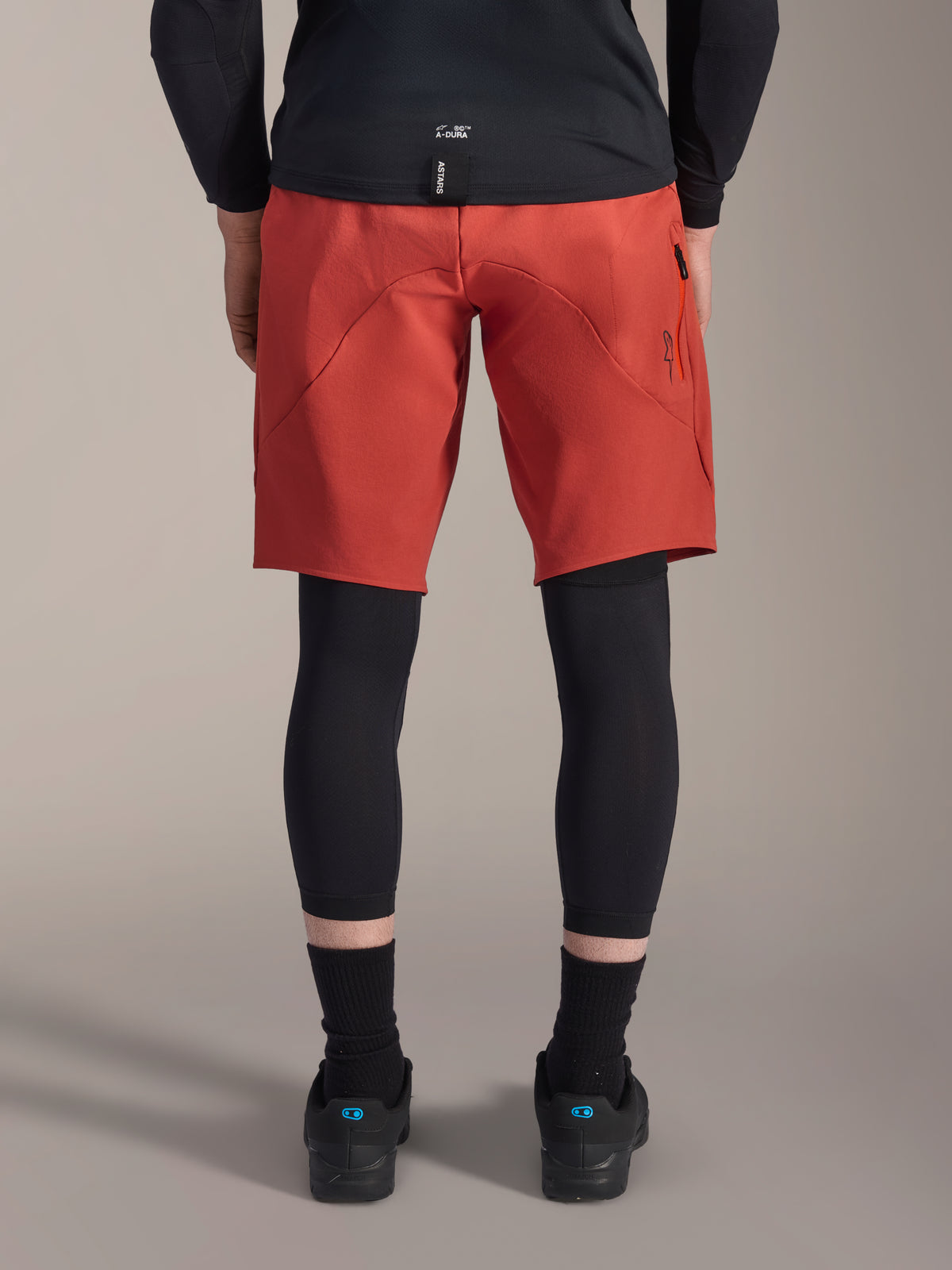 Alpinestars A-Motion Plasma Knee Sleeve, black MTB knee protectors, back view worn under red mountain bike shorts with a black jersey and technical cycling shoes, protective gear for mountain biking