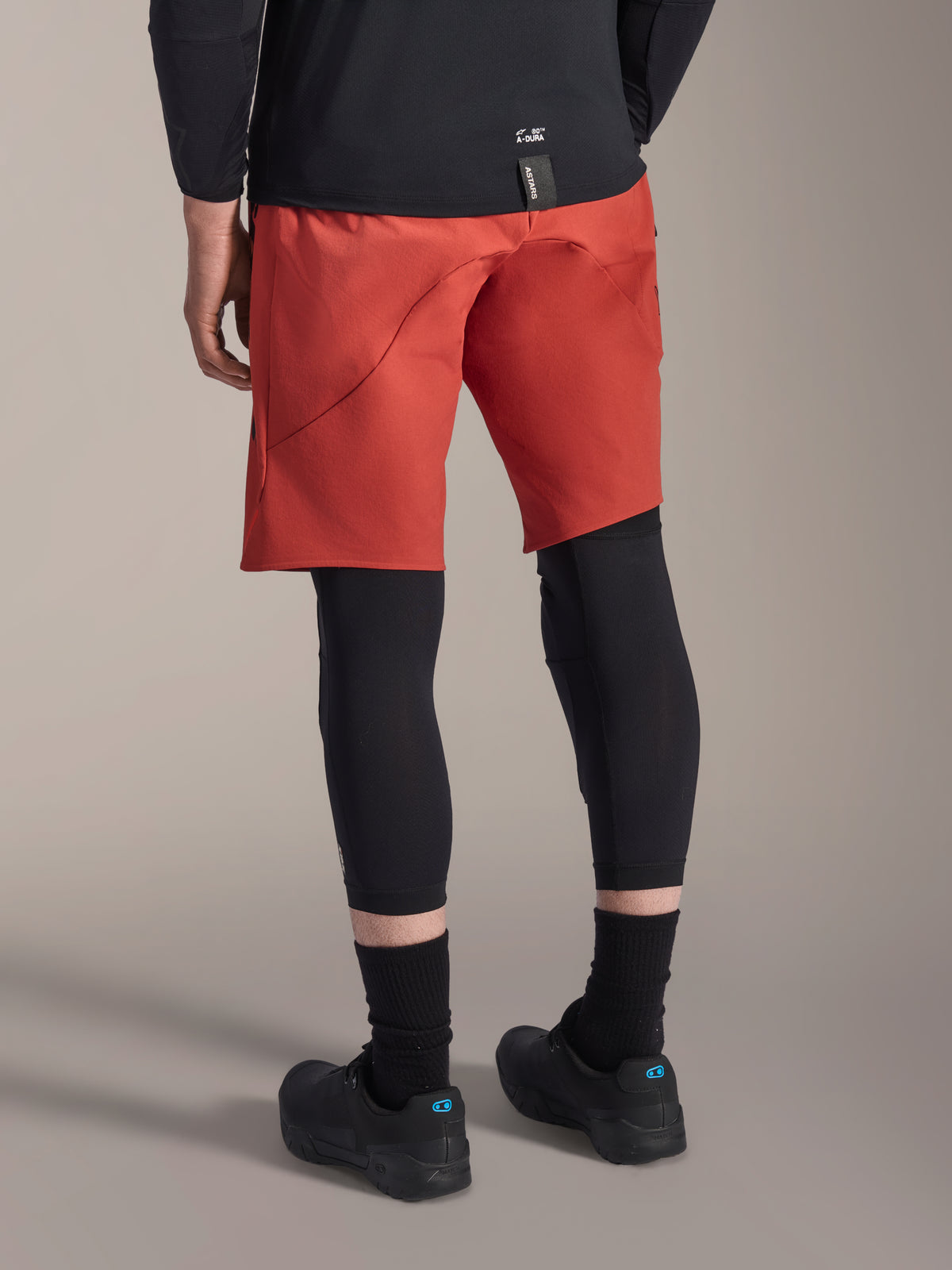 Alpinestars A-Motion Plasma Knee Sleeve, Black MTB Knee Protector, worn with red mountain bike shorts and black riding shoes, rear view showing the low-profile slip-on design for off-road cycling