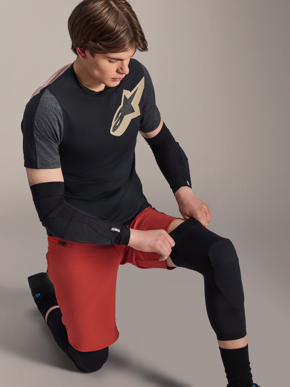 Alpinestars A-Motion Plasma Knee Sleeve, MTB Knee Protector, Black, worn by a rider in a kneeling position, paired with a black and grey t-shirt featuring a gold logo, red shorts, and black elbow guards, professional mountain bike protective equipment