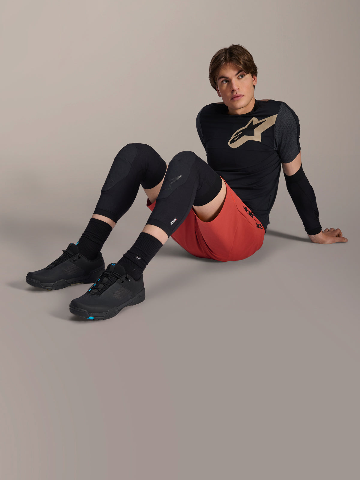 Alpinestars A-Motion Plasma Knee Sleeve, black mountain bike knee protectors, worn by a male model sitting in complete cycling gear with red shorts and black performance tee, showcasing the low-profile protective design.