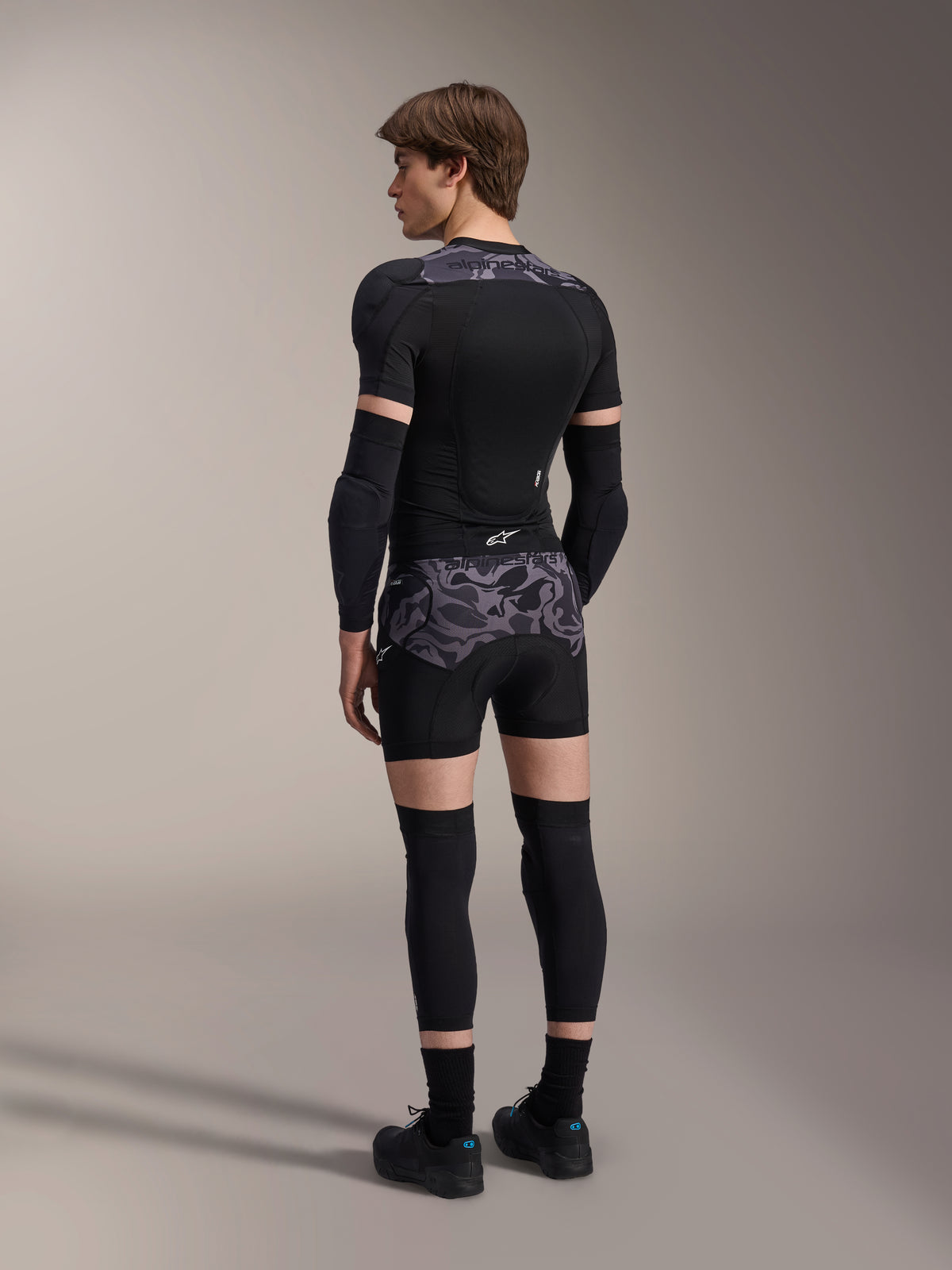 Model w bokserkach ochronnych Alpinestars A-Motion Plasma w kolorze Black Camo i pasującej kurtce ochronnej A-Motion Plasma, widok z tyłu, szary wzór kamuflażu, ochraniacze łokci, ochraniacze kolan, buty rowerowe, kompletny zestaw ochronny MTB