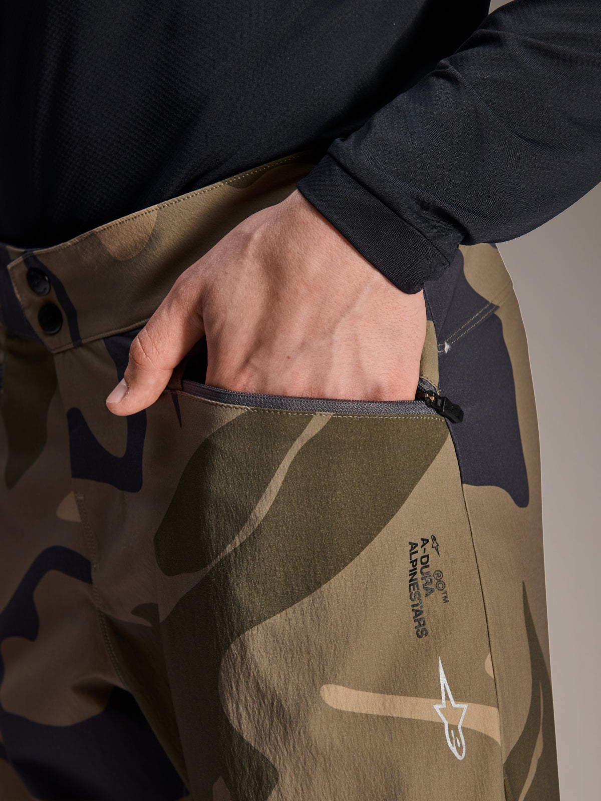 Close-up detail of Alpinestars A-Dura Camo Pant, Brown Camouflage Mountain Bike Pants, featuring a hand reaching into a zippered pocket, double snap waist closure, and Alpinestars branding on the thigh.