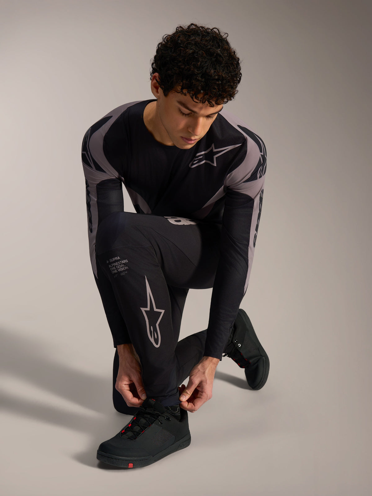 Czarne spodnie Alpinestars A-Supra Elite z szarymi logotypami, noszone przez klęczącego modela do jazdy MTB.