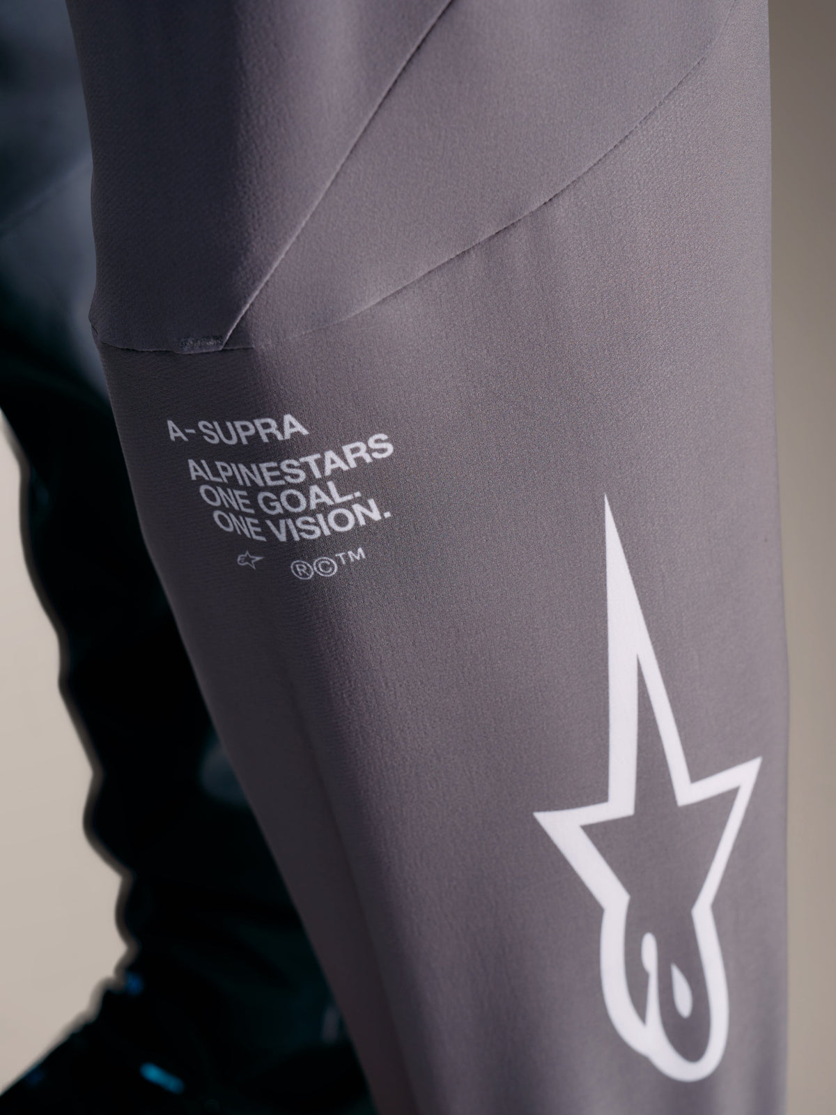 Spodnie A-Supra Elite, szare, z białym brandingiem Alpinestars i mottem wyścigowym „One Goal. One Vision.”