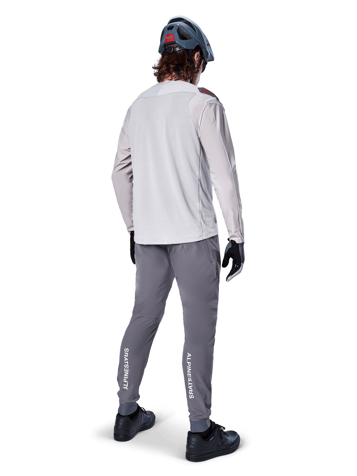 Alpinestars A-Aria Elite Pants, spodnie rowerowe MTB, ciemnoszare, widok z tyłu na rowerzystę w pełnym stroju, z białym logo Alpinestars na dolnej części nogawek, sportowy krój z laserowymi perforacjami, zestawione z jasnoszarą koszulką z długim rękawem i szarym kaskiem MTB