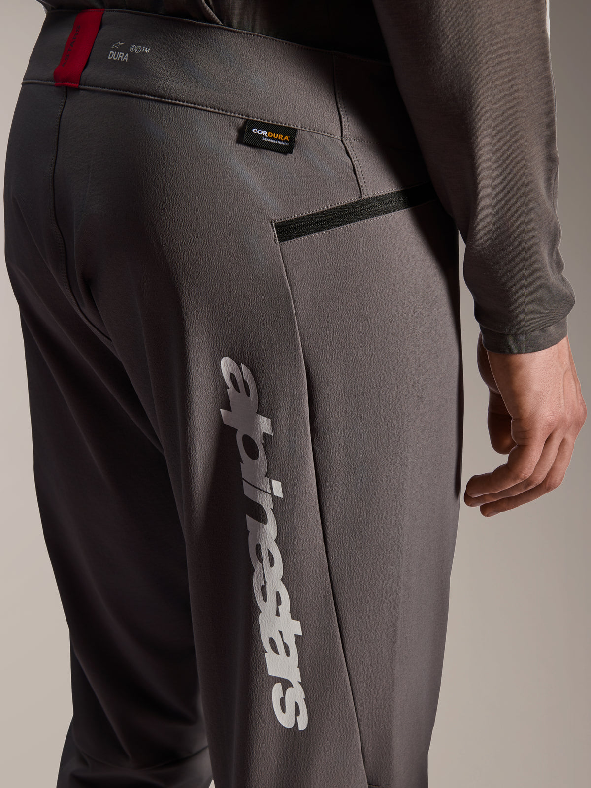 Alpinestars A-Dura Elite Pants, Dark Gray MTB Pants, close-up view of hip and side thigh, featuring large white vertical Alpinestars logo, Cordura fabric tag, zippered pocket, and red accent on the waistband