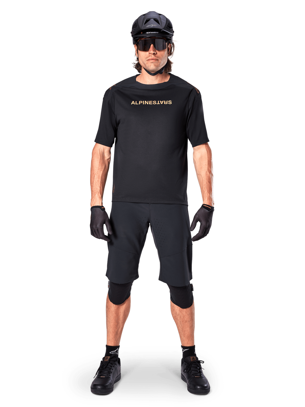 Męski rowerzysta górski w czarnych spodenkach Alpinestars A-Aria Elite, w połączeniu z pasującą czarną koszulką Alpinestars z krótkim rękawem, czarnym kaskiem, rękawicami i ochraniaczami na kolana, kompletny strój rowerowy off-road