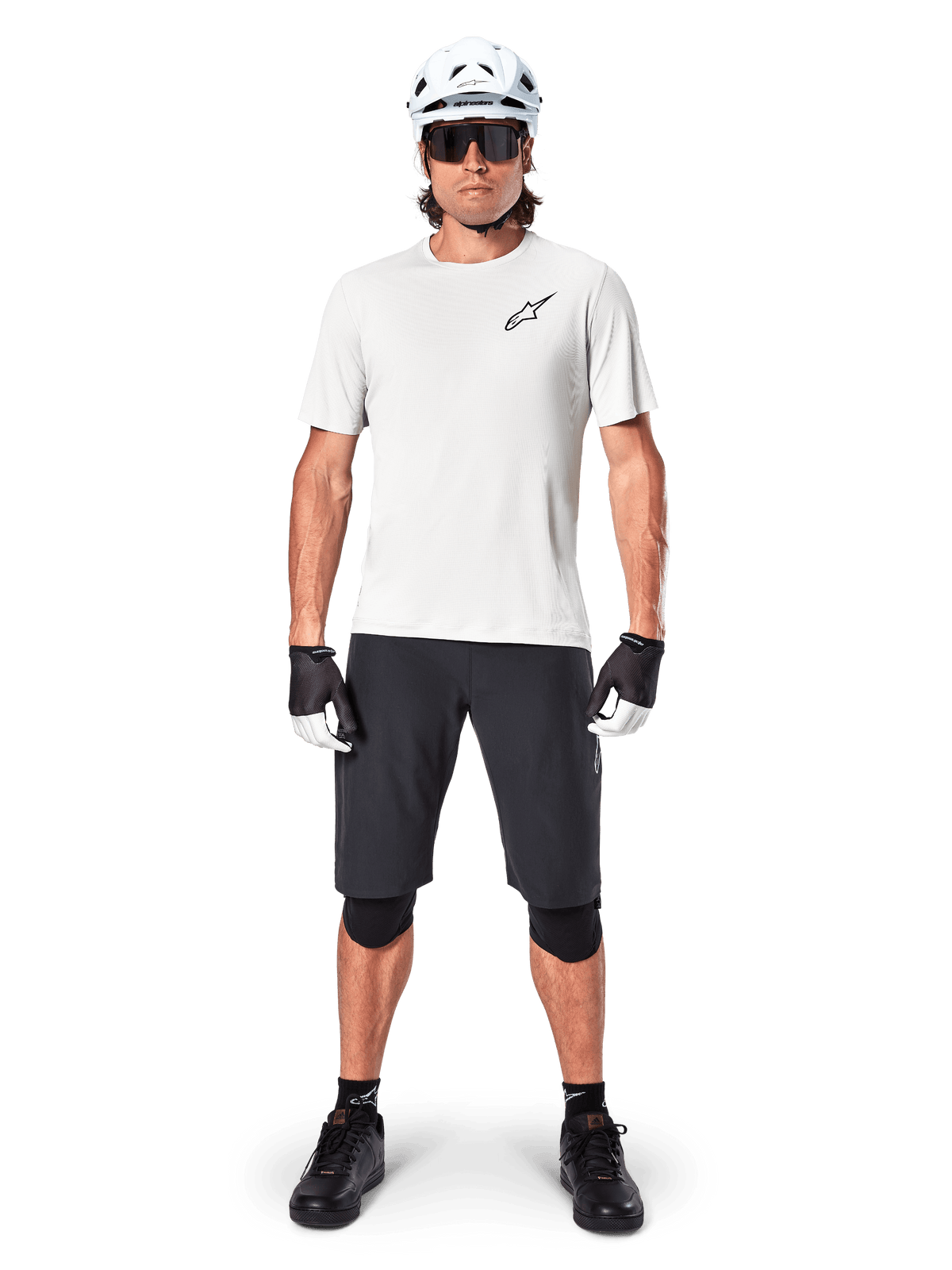 Rowerzysta górski w czarnych spodenkach Alpinestars A-Aria, biała koszulka techniczna, biały kask i czarne ochraniacze na kolana, profesjonalny strój do jazdy terenowej, widok z przodu całej sylwetki