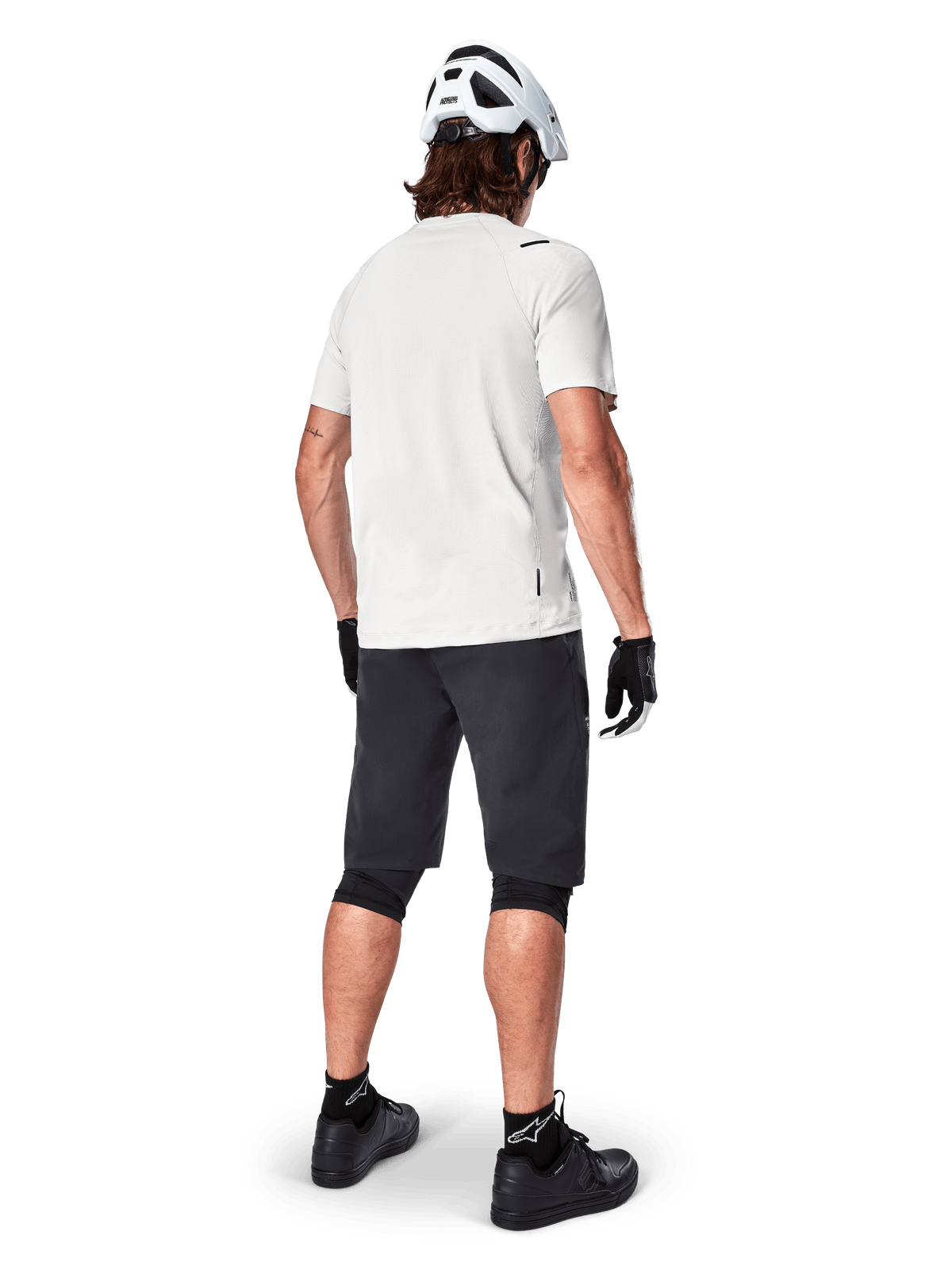 Rowerzysta górski widoczny od tyłu, ubrany w szorty Alpinestars A-Aria w kolorze czarnym, białą techniczną koszulkę z krótkim rękawem, biały kask, czarne rękawiczki i czarne buty MTB, odzież rowerowa do jazdy po szlakach