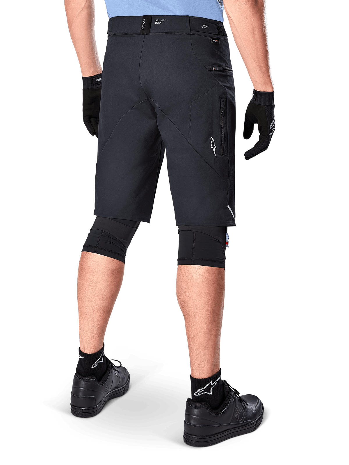 Alpinestars A-Dura Elite Shorts, czarne, widok od tyłu rowerzysty w stroju MTB, w tym czarne nakolanniki, rękawiczki i skarpetki z białym logo, boczne kieszenie na zamek i odblaskowe akcenty, wytrzymały materiał Cordura