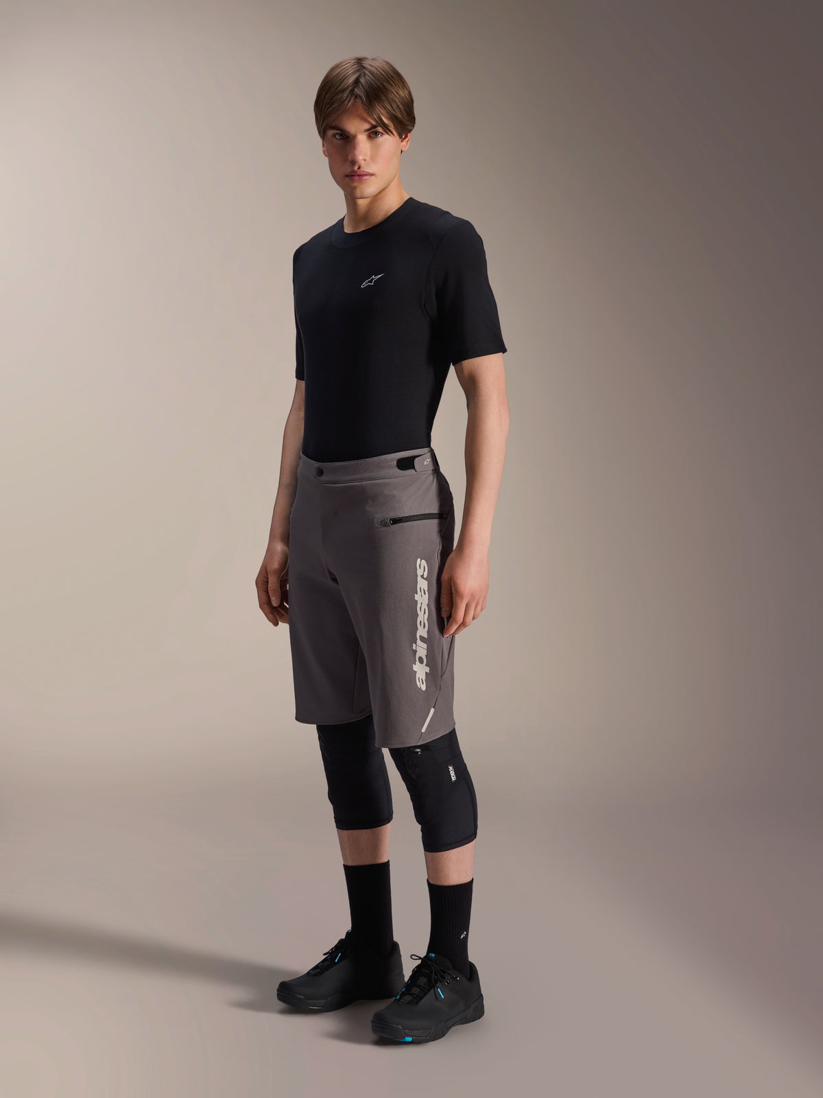 Spodenki Alpinestars A-Dura Elite, ciemnoszare spodenki rowerowe MTB, prezentowane przez modela w czarnym t-shircie i ochraniaczach na kolana, z pionowym białym logo Alpinestars na lewej nogawce, techniczny materiał Cordura do jazdy w terenie.