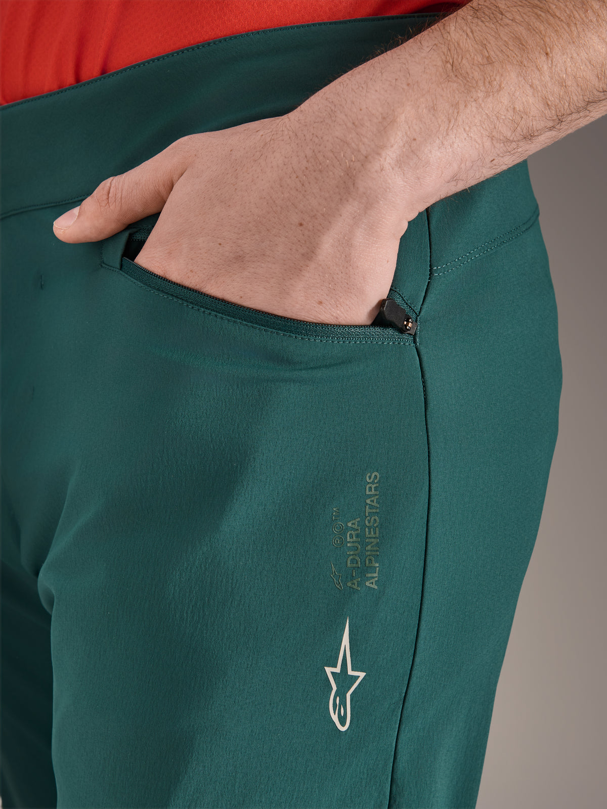 Spodnie Alpinestars A-Dura w kolorze Dark Pine, zbliżenie na kieszeń zapinaną na zamek z dłonią, białe logo i branding Alpinestars na udzie, wytrzymałe spodnie rowerowe MTB
