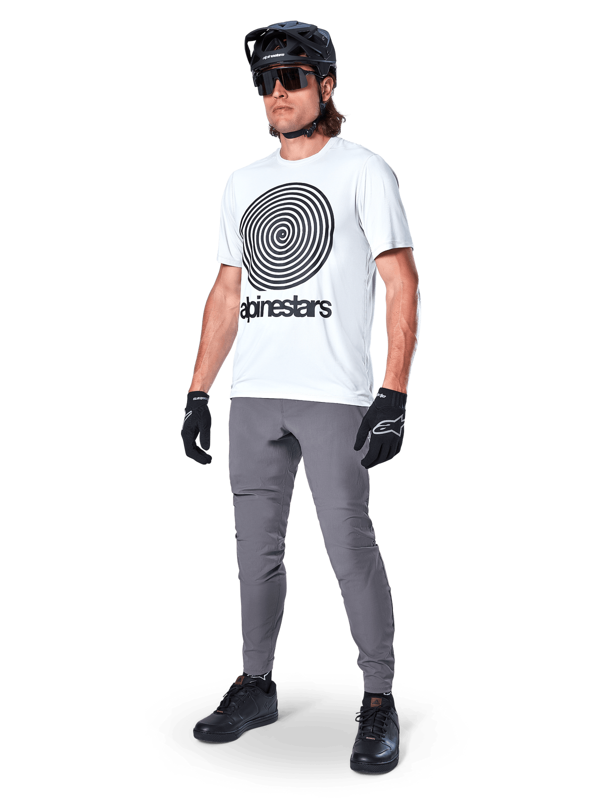 Alpinestars A-Dura Pants, spodnie MTB, ciemnoszare, noszone z białym t-shirtem z grafiką spirali, czarnym kaskiem MTB, okularami przeciwsłonecznymi i czarnymi rękawicami, wytrzymały materiał rozciągliwy w 2 kierunkach do jazdy w terenie