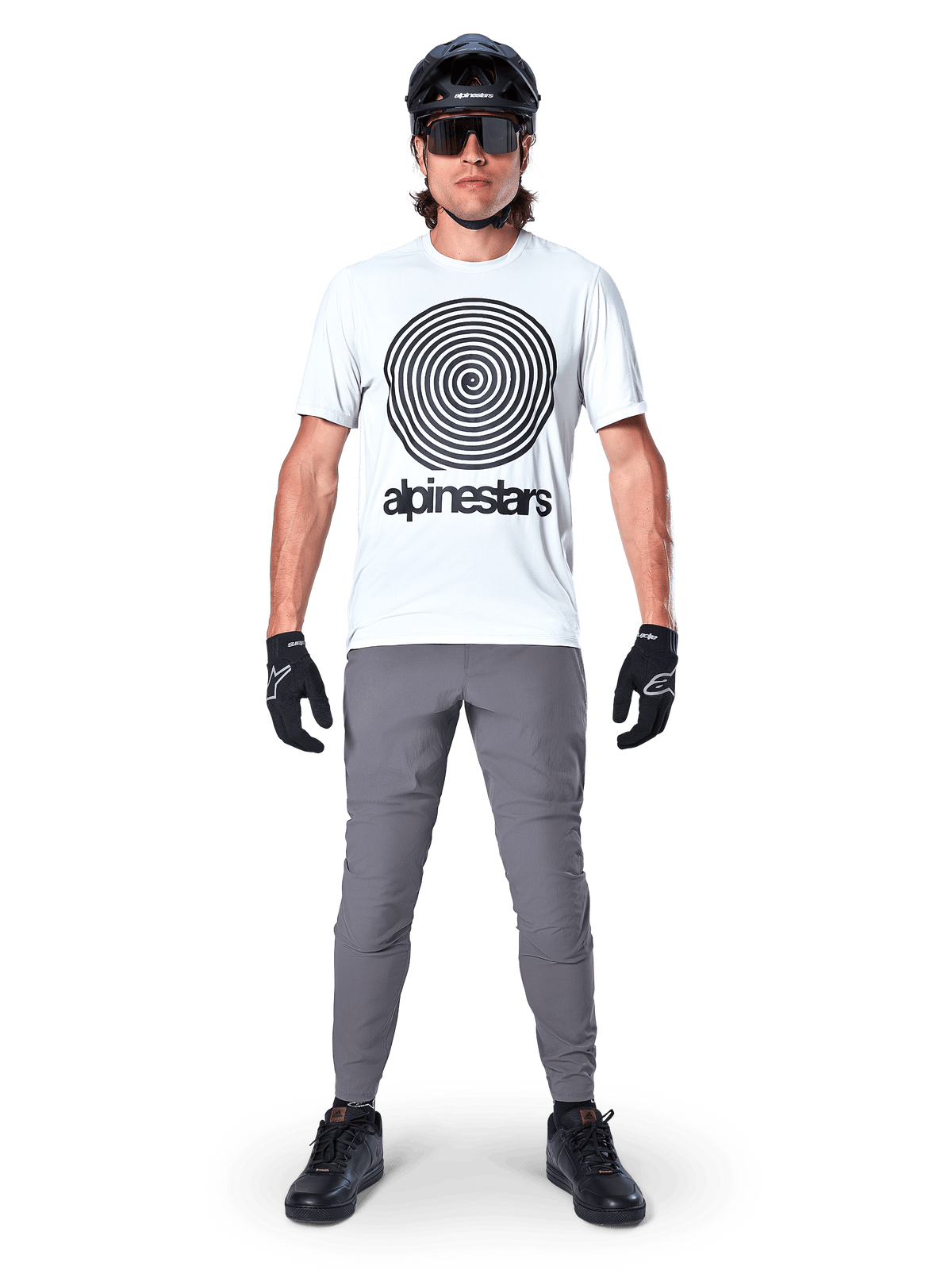 Alpinestars A-Dura Pants, rowerowe spodnie MTB, ciemnoszare, zestawione z białym t-shirtem z grafiką, czarnym kaskiem i rękawiczkami, charakteryzują się trwałą konstrukcją z dwukierunkowego stretchu i regularnym krojem do jazdy na rowerze górskim