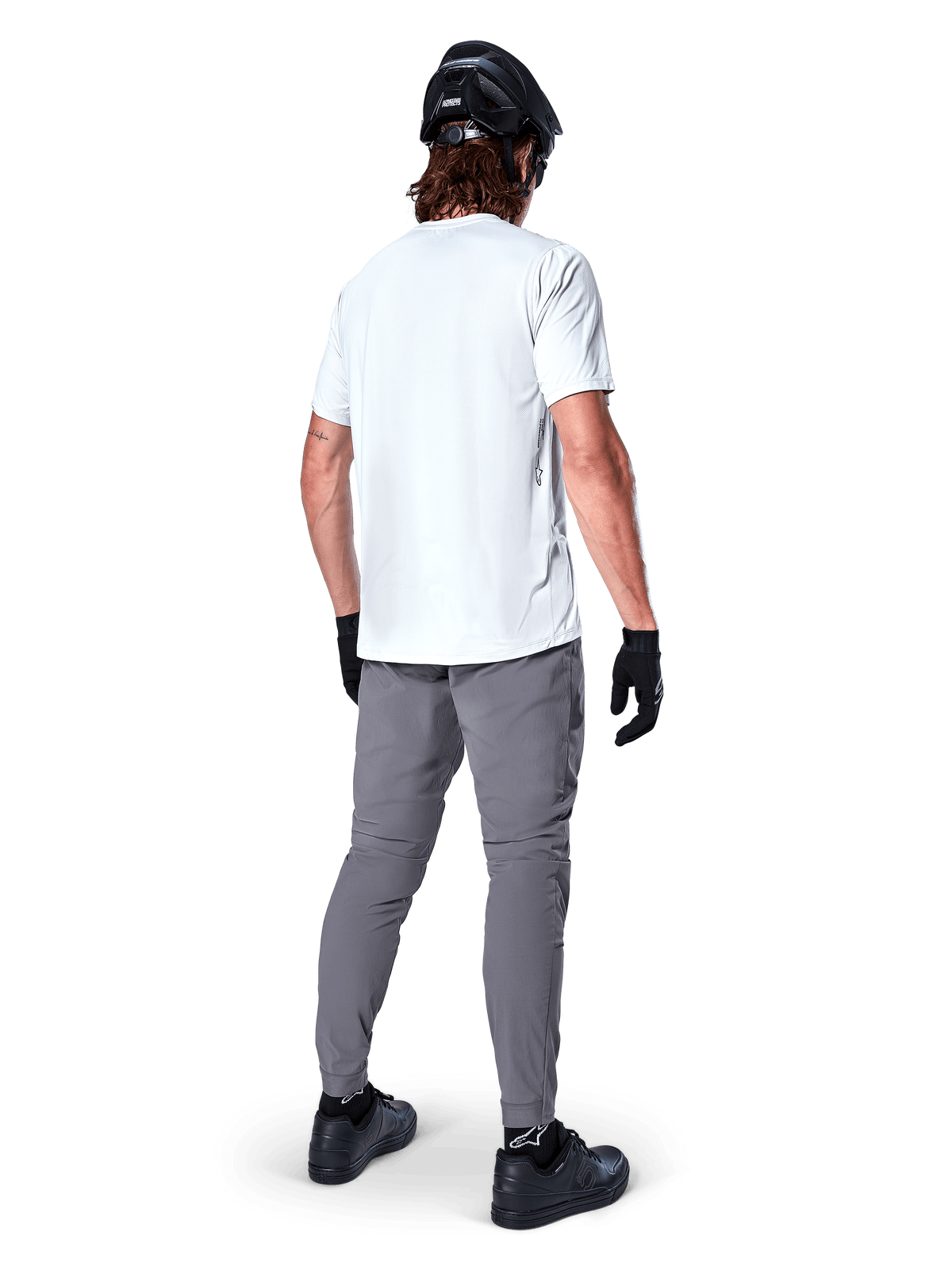 Alpinestars A-Dura Pants, ciemnoszare spodnie na rower górski widoczne z tyłu, zestawione z białym t-shirtem z krótkim rękawem, czarnym kaskiem MTB, czarnymi rękawiczkami i butami, krój regular fit z elastycznymi mankietami, wytrzymała odzież do jazdy w terenie