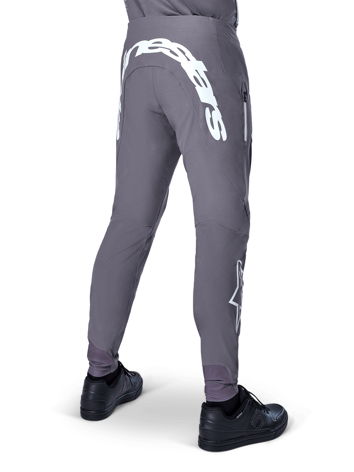 Alpinestars A-Supra Race Pants, spodnie rowerowe MTB, ciemnoszare, widok z tyłu, czarne buty, duży biały napis Alpinestars na karczku i logo gwiazdy na nogawce, kieszeń zapinana na zamek, elastyczny materiał techniczny do wyścigów MTB