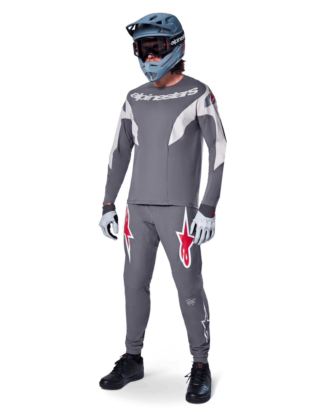 Alpinestars A-Supra Race Pants w kolorze ciemnoszarym, z wyraźnymi czerwonymi i białymi logotypami na udach, noszone przez zawodnika jako część kompletnego zestawu wyścigowego MTB z pasującą szarą koszulką, niebiesko-szarym kaskiem full-face i jasnoszarymi rękawicami, profesjonalna odzież do wyścigów MTB