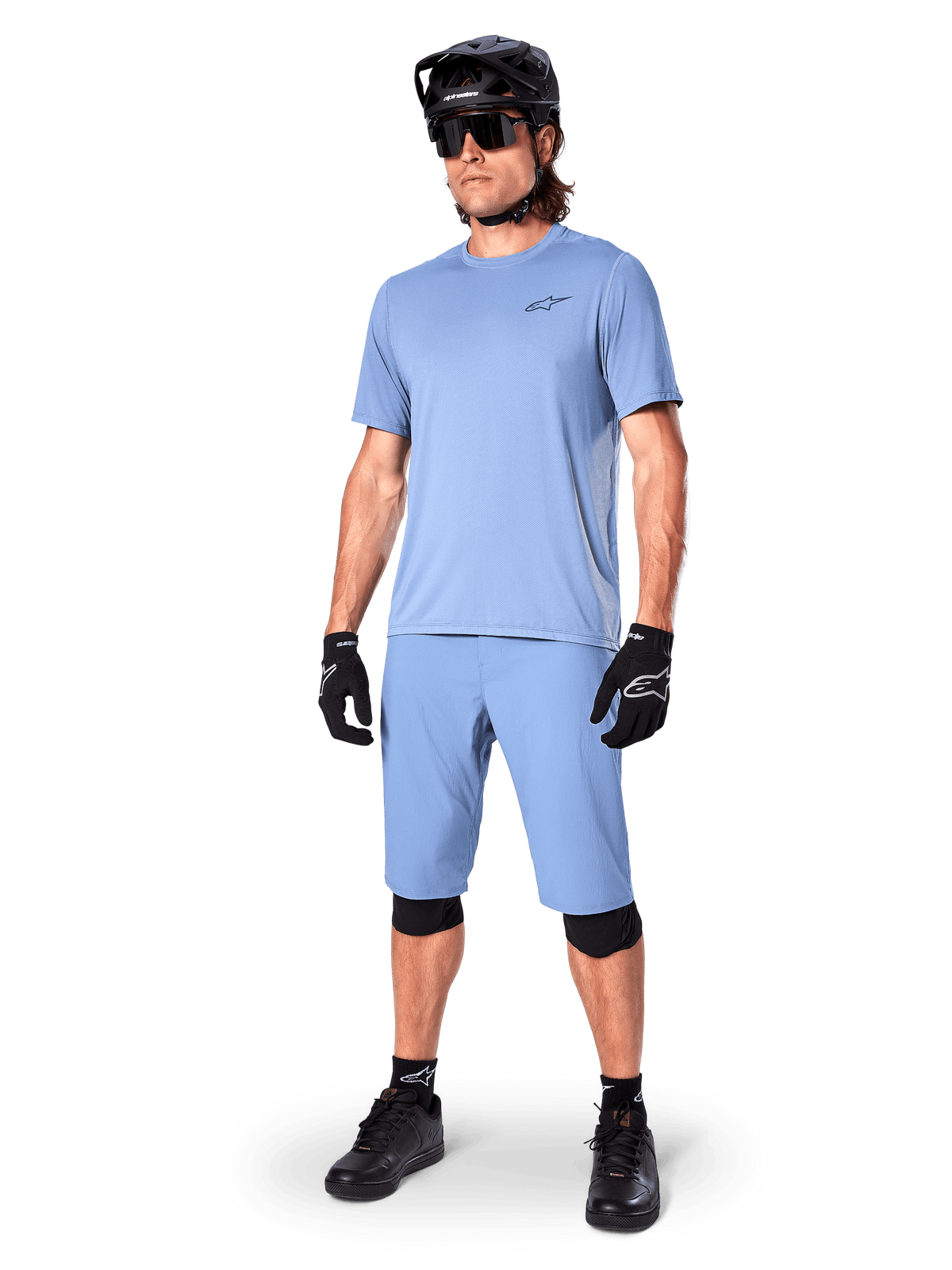 Model ubrany w spodenki Alpinestars A-Dura w kolorze Infinity Blue, pasującą jasnoniebieską koszulkę z krótkim rękawem, czarny kask MTB, okulary przeciwsłoneczne i czarne rękawiczki, kompletny strój do kolarstwa górskiego do jazdy trailowej