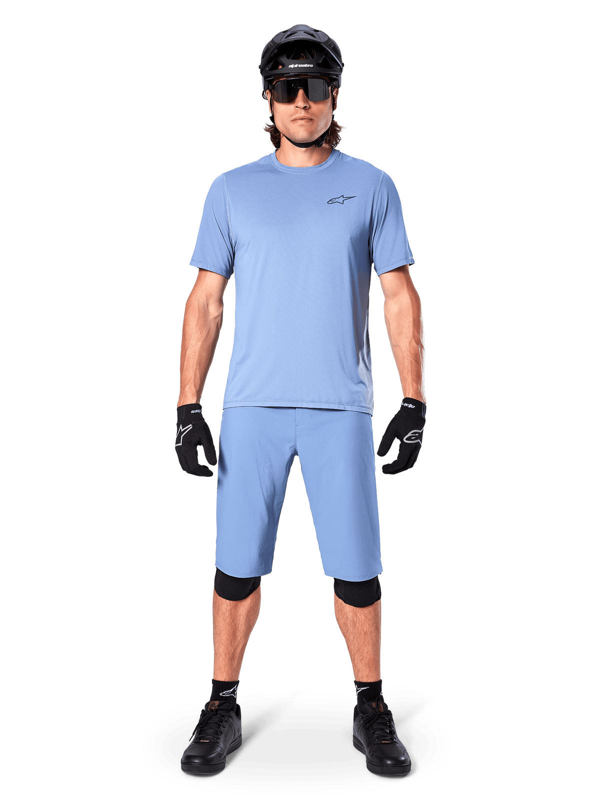 Model w spodenkach Alpinestars A-Dura w kolorze Infinity Blue, pasującej jasnoniebieskiej koszulce, czarnym kasku MTB, czarnych okularach przeciwsłonecznych, czarnych rękawicach i nakolannikach, kompletny strój MTB do jazdy po szlakach