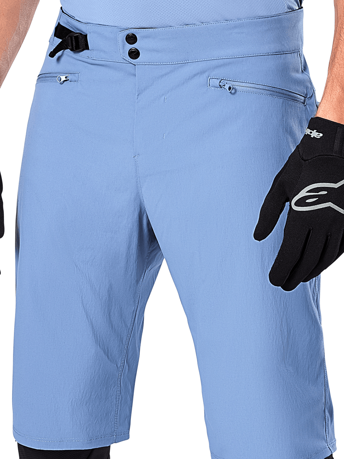 Alpinestars A-Dura Shorts, niebieskie spodenki MTB (Infinity Blue), zbliżenie na talię z podwójnym zapięciem na napy, regulowanym bocznym ściągaczem i kieszeniami na zamek z podszewką z siatki, widoczne z czarną rękawiczką Alpinestars